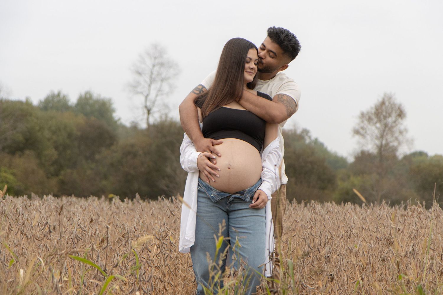 A man hugging his pregnant wife from behind while they stand in a field.
