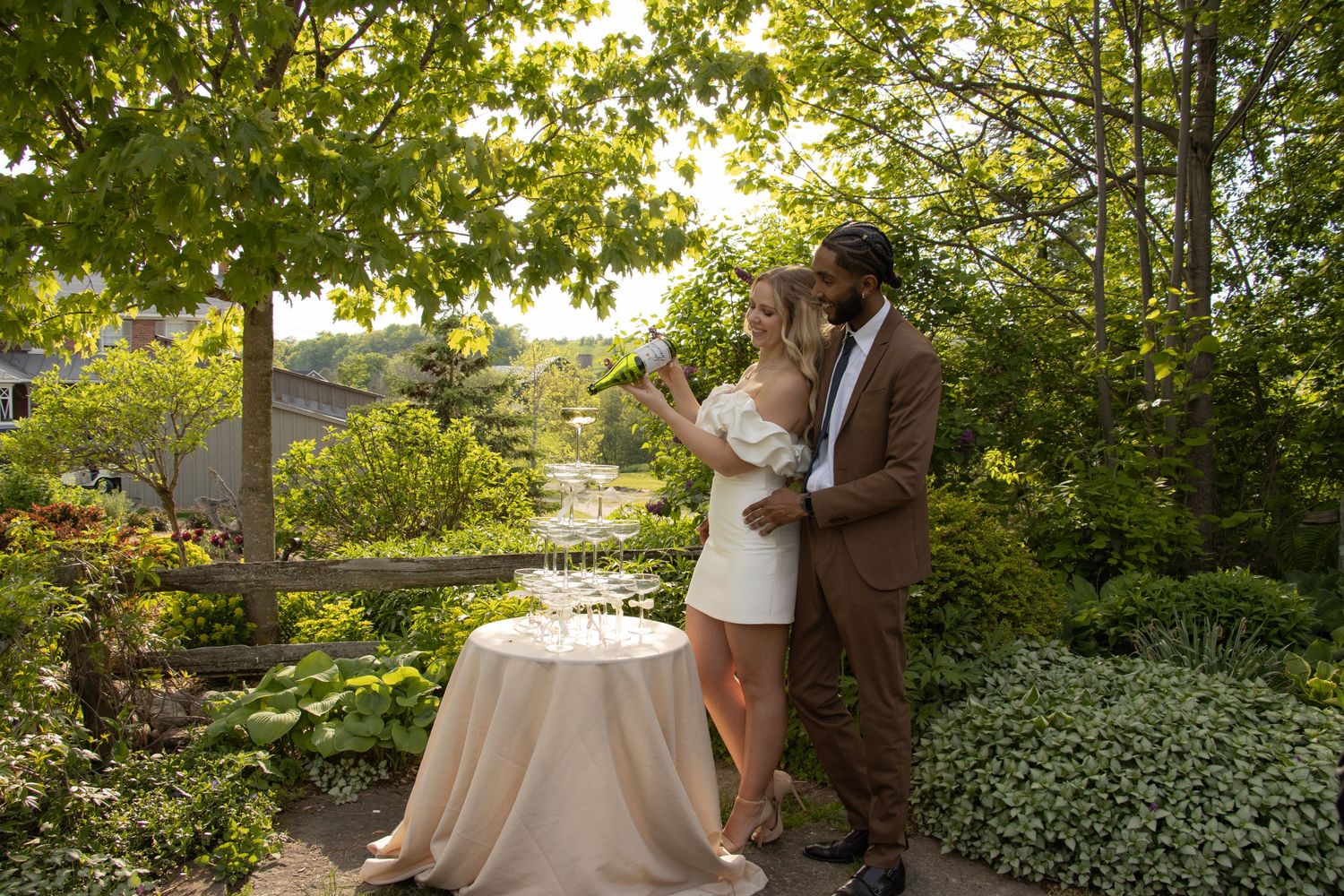 A couple stands outdoors in a lush garden. The woman, dressed in a white dress, is pouring champagne into a tower of gla