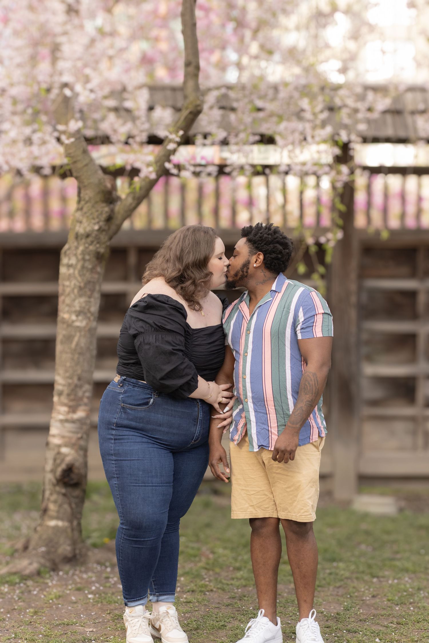 A couple stands outdoors, kissing under a blossoming cherry tree. The woman wears an off-shoulder black top and jeans, w
