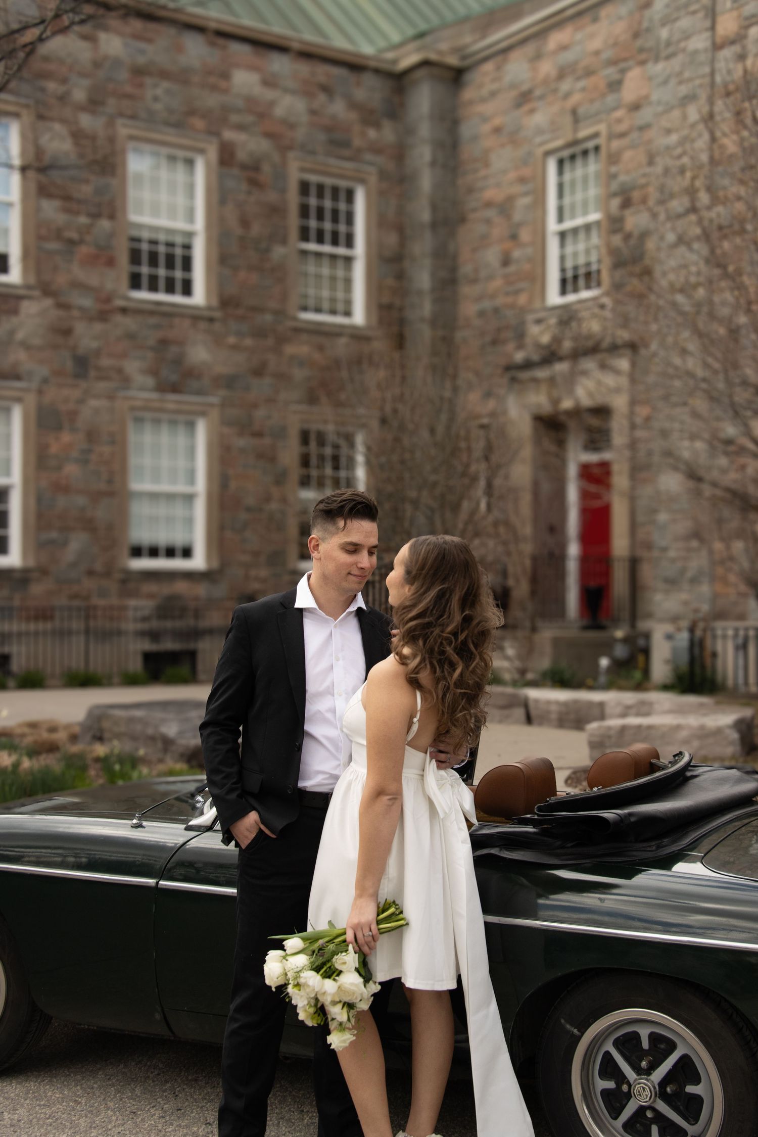 A bride and groom beside a convertible car parked in front of a stone building. The man is in a suit, and the woman is i