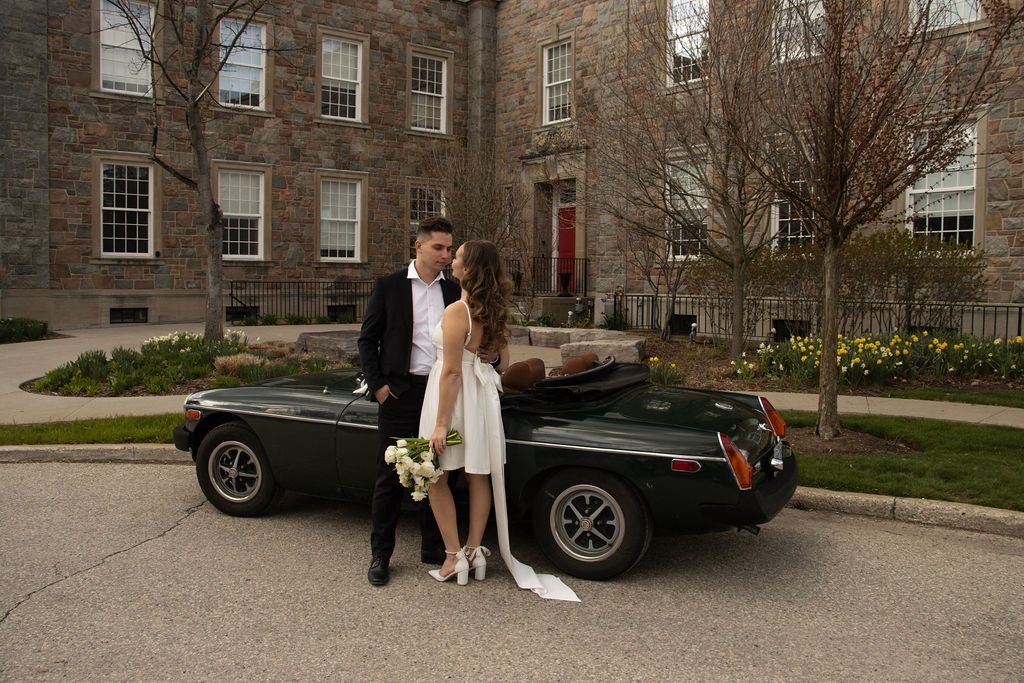 A bride and groom beside a convertible car parked in front of a stone building. The man is in a suit, and the woman is i