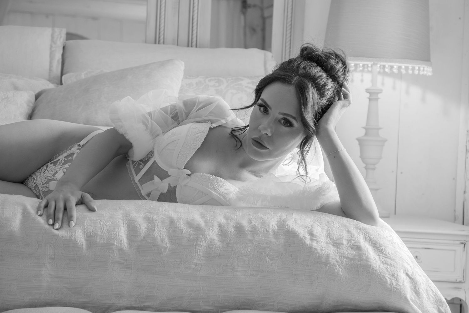 A woman in elegant lingerie reclines on a bed in a softly lit room. She is wearing a sheer top over a lacy bra and match