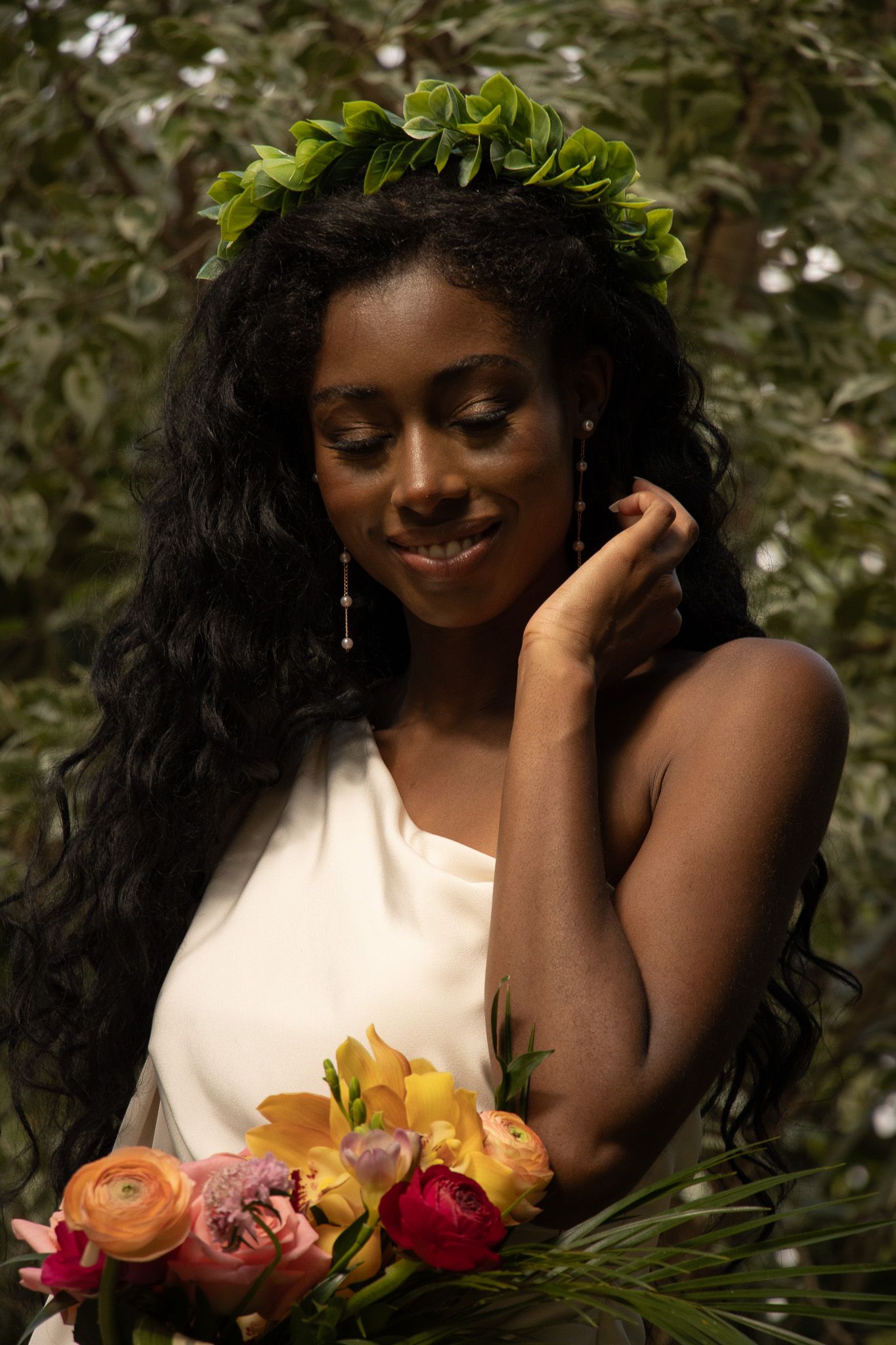 A woman dressed in a white one-shoulder outfit stands outdoors, smiling gently with her eyes closed. She wears a leafy g