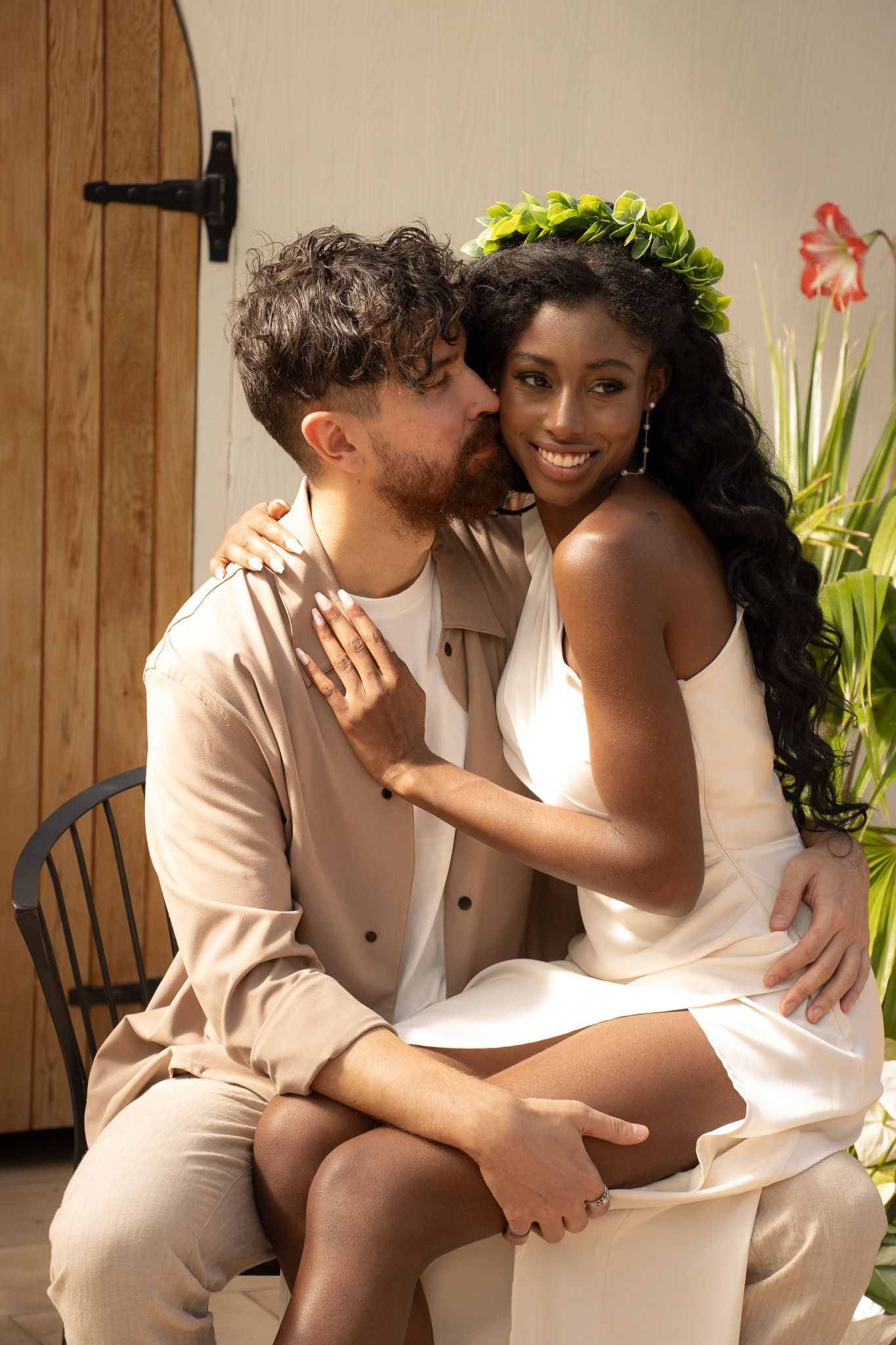 A man with dark hair and a beard is seated on a chair, embracing and kissing the cheek of a woman with long, curly hair
