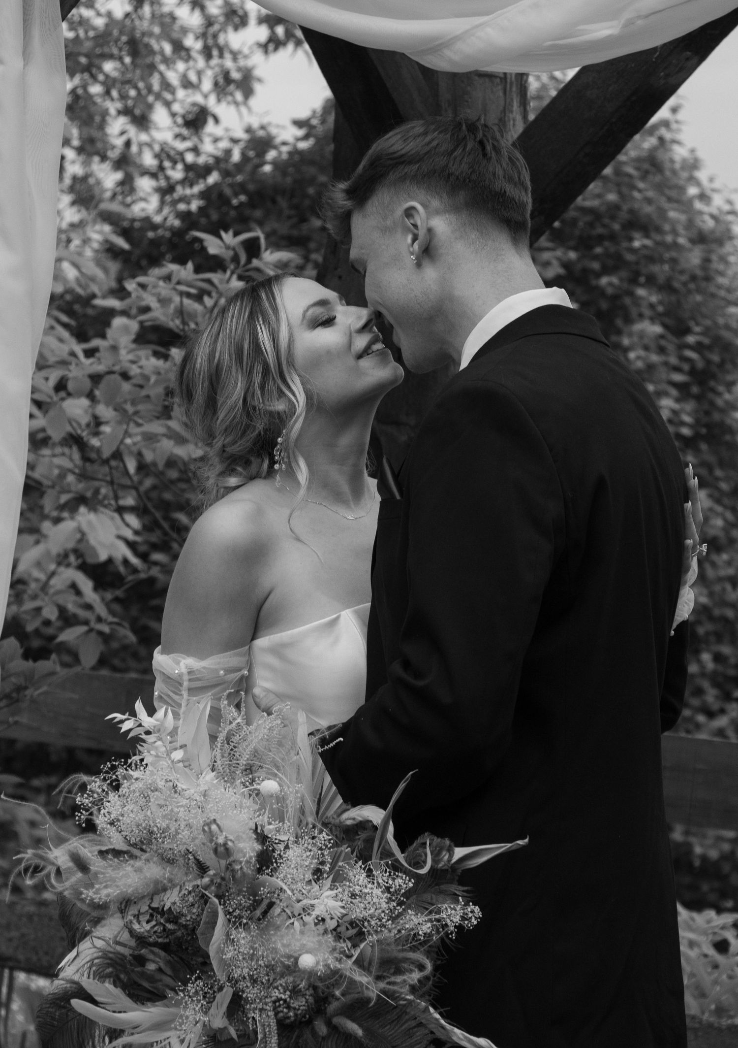 A bride and groom lean in for a kiss on their wedding day. The bride, in an off-shoulder white gown, holds a bouquet of