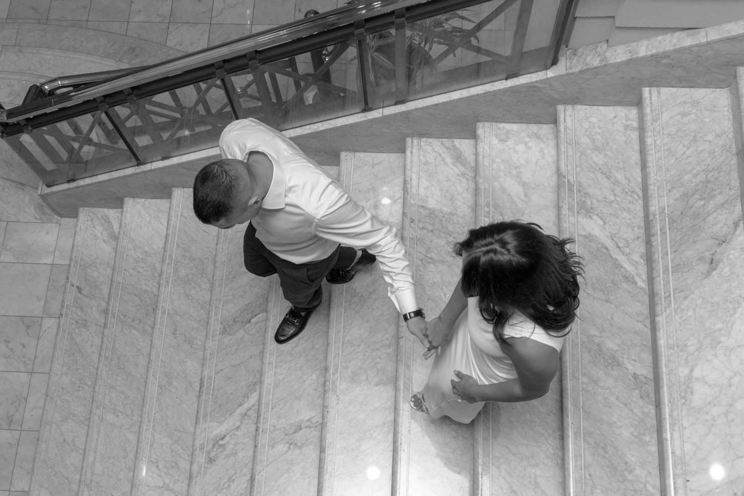 A black-and-white photo of a couple holding hands while walking up a wide staircase. The woman is wearing a long, elegan