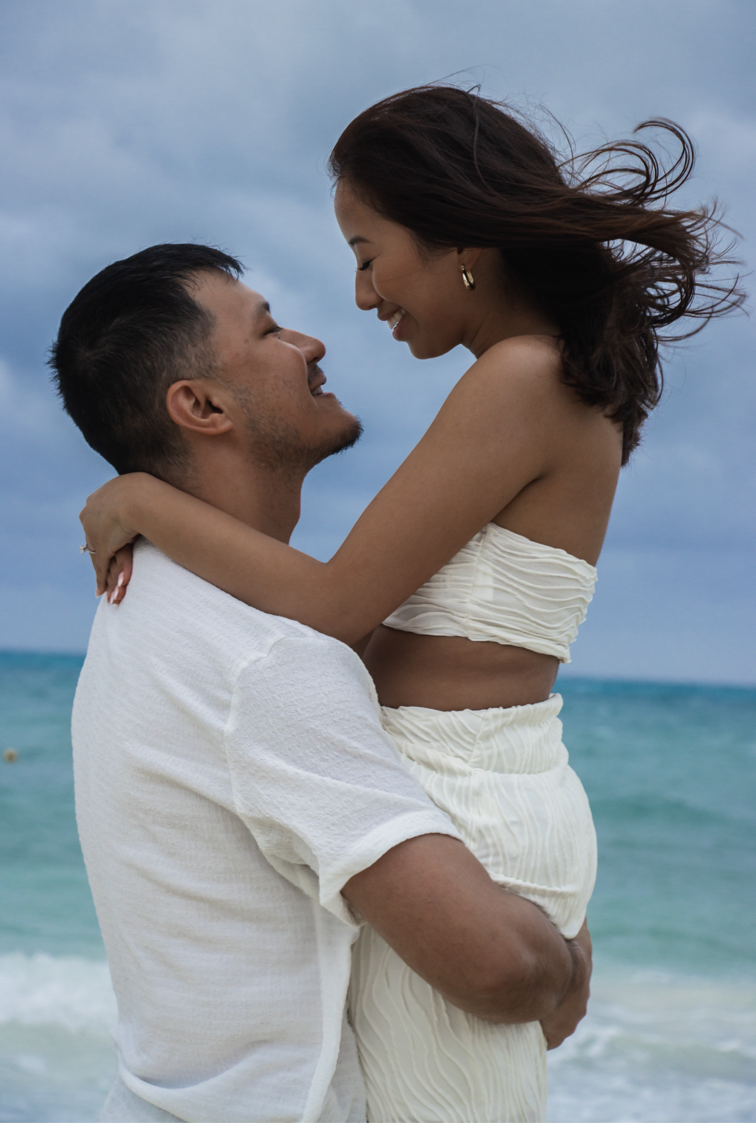 A couple embraces by the ocean, with the man lifting the woman in his arms. They share a joyful moment, looking into eac