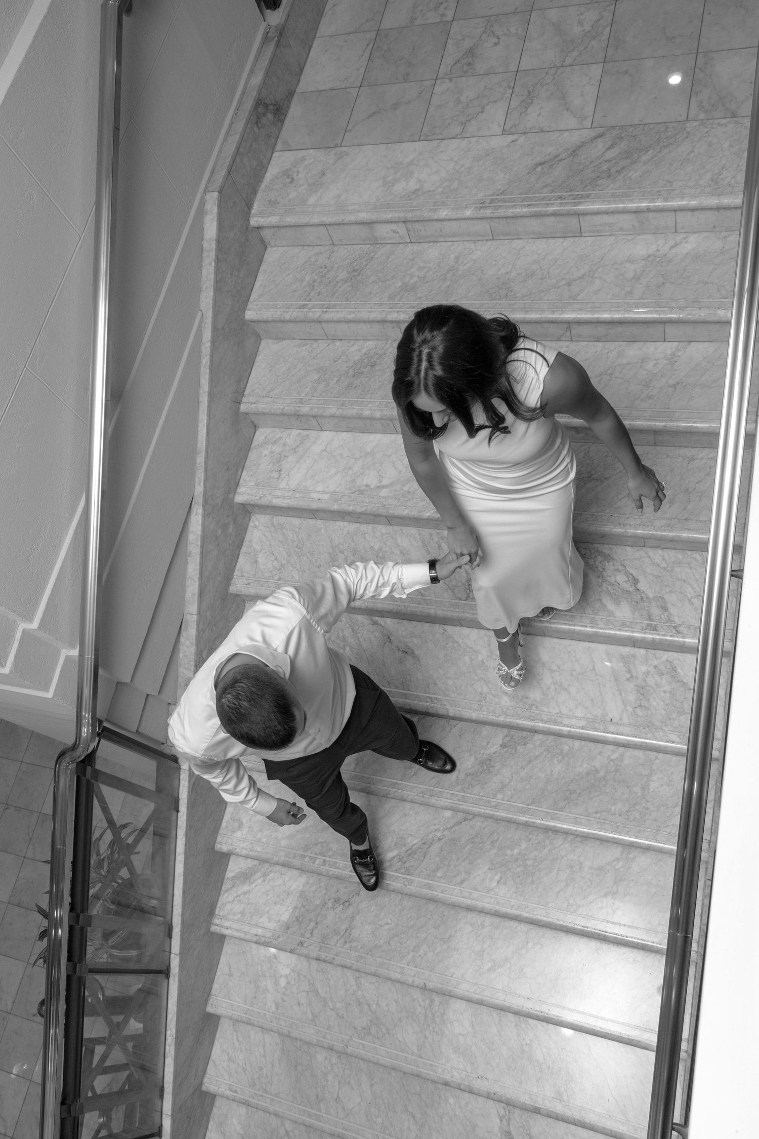 A black-and-white photo of a couple holding hands while walking up a wide staircase. The woman is wearing a long, elegan