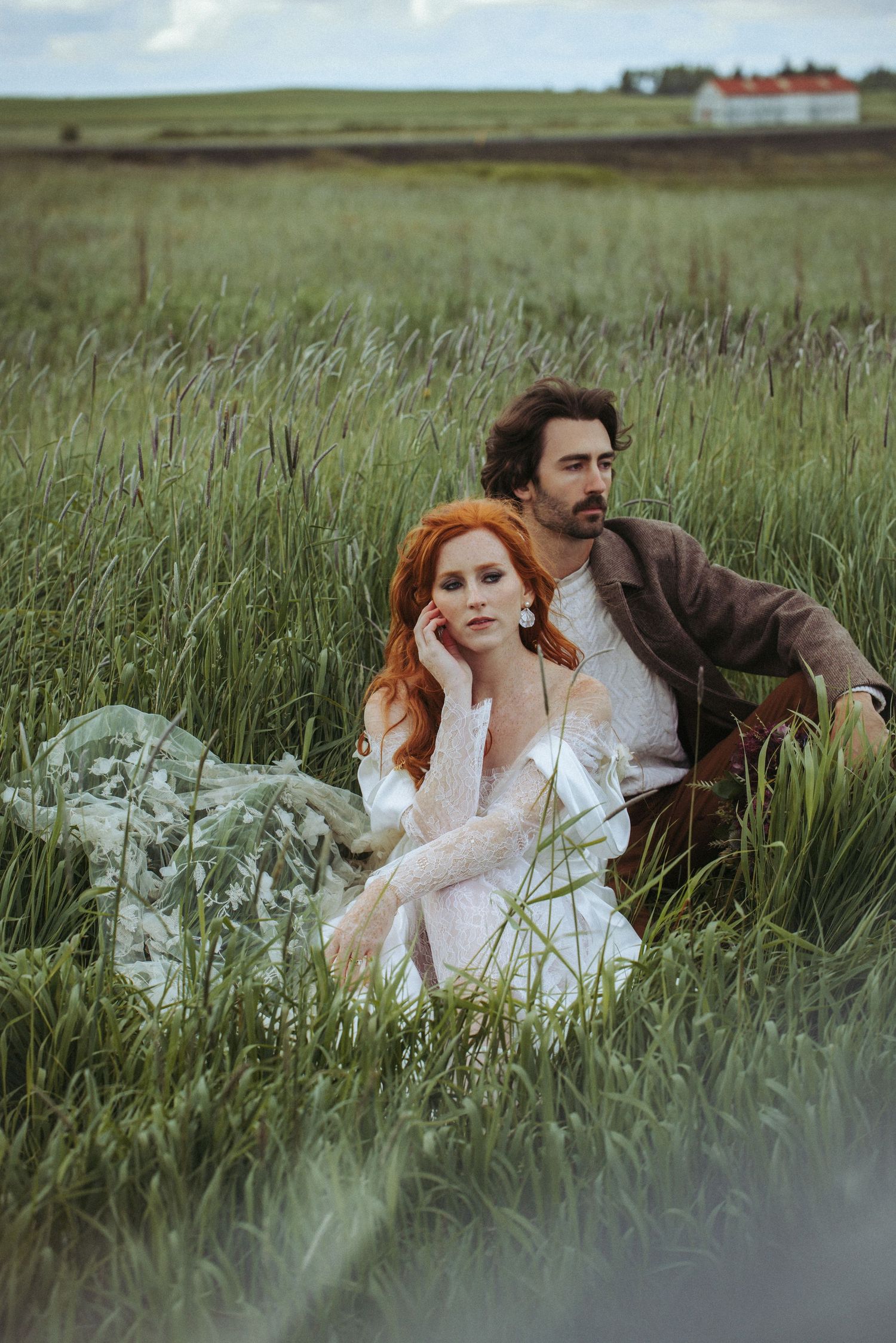 A couple sits together in a field of tall grass at dusk, with the person wearing a romantic white lace dress in foreground.