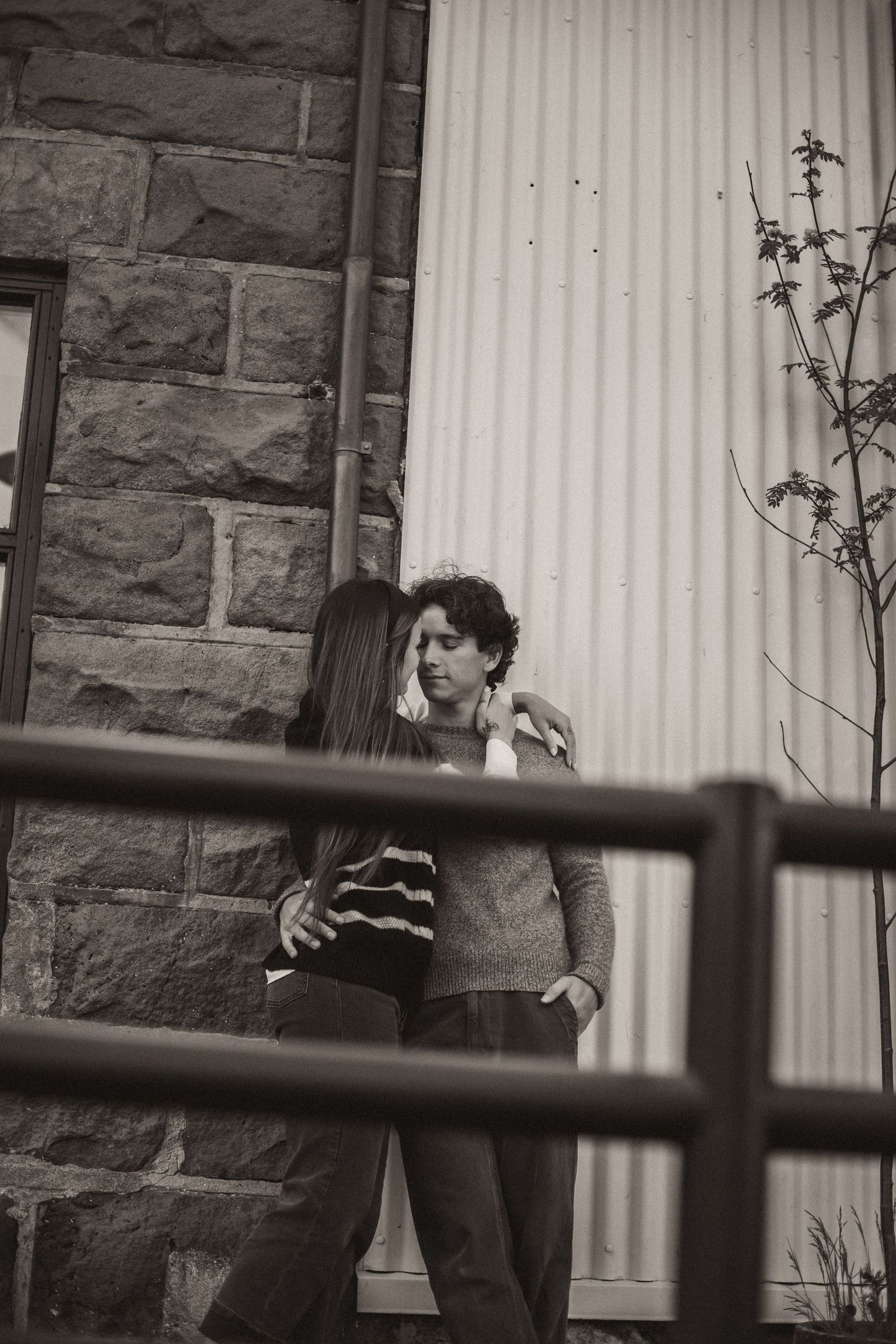 A monochrome image captures figures through wooden fence posts against a stone wall.