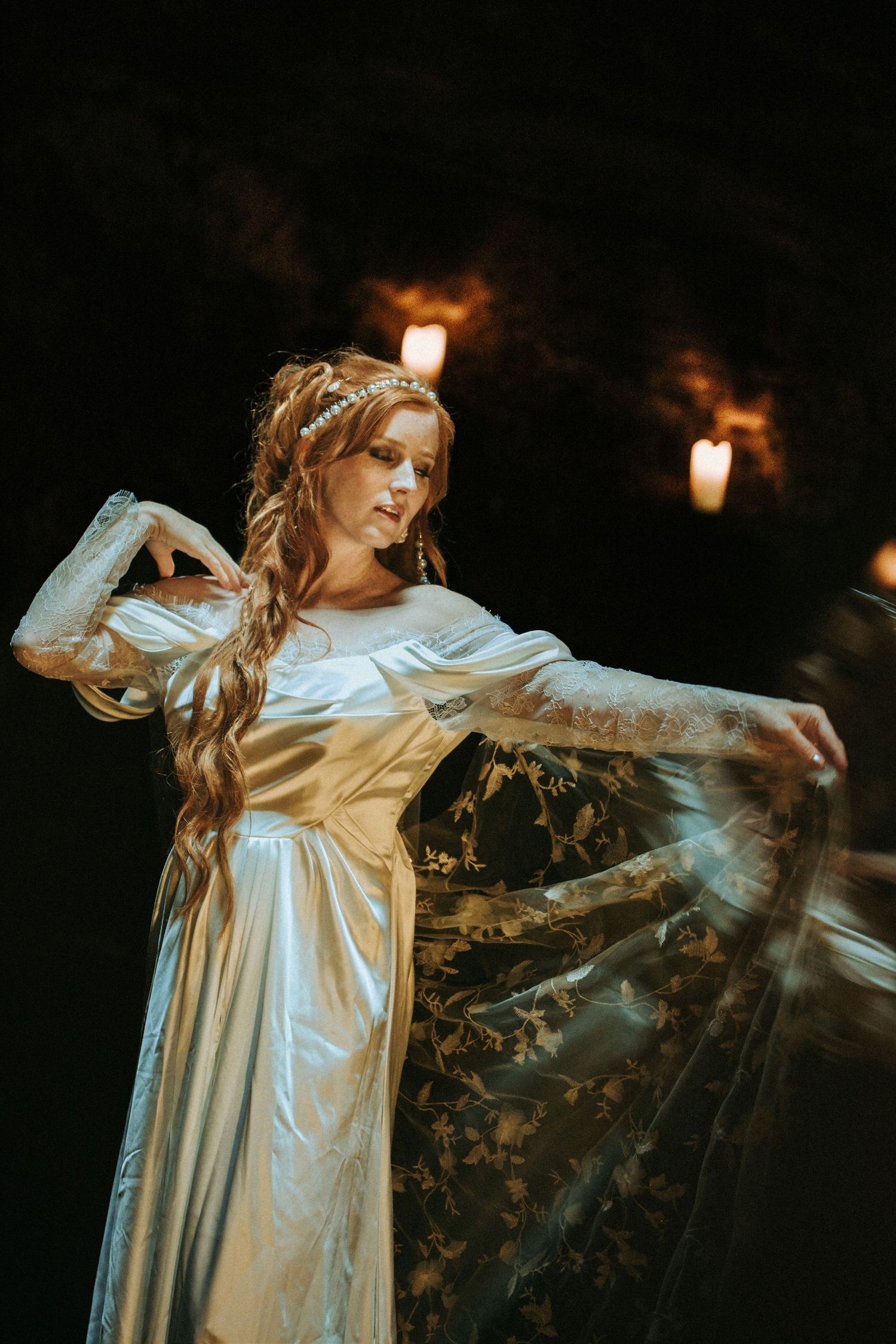 A person in a flowing white vintage dress poses dramatically in a dark room lit by candlelight.