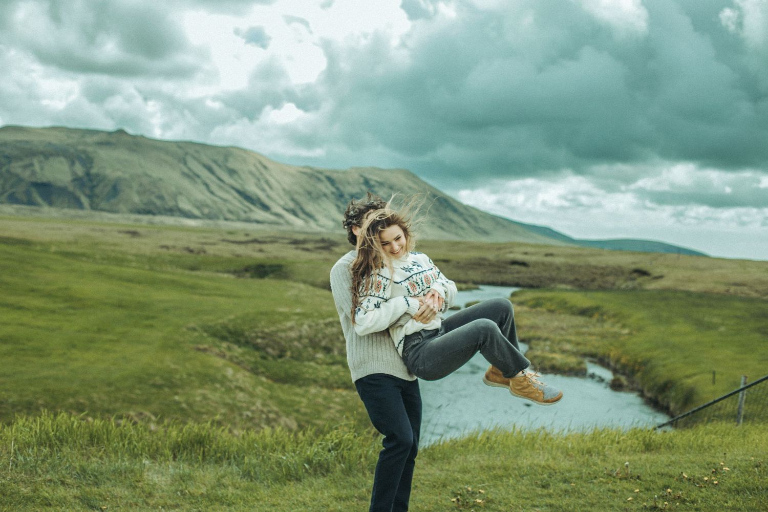 Person being lifted playfully in mountain landscape with river and cloudy sky while wearing cozy sweaters..