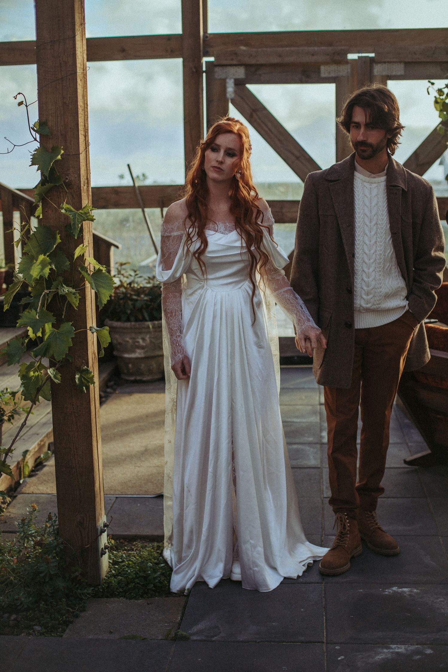 A couple poses in a greenhouse setting with the bride wearing a flowing white off-shoulder dress and groom in brown suit.
