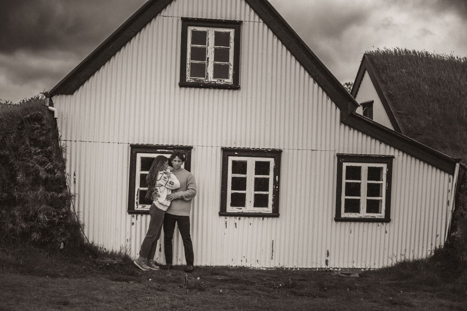 Black and white photo of a couple standing by a historic white wooden house with windows on a cloudy day.