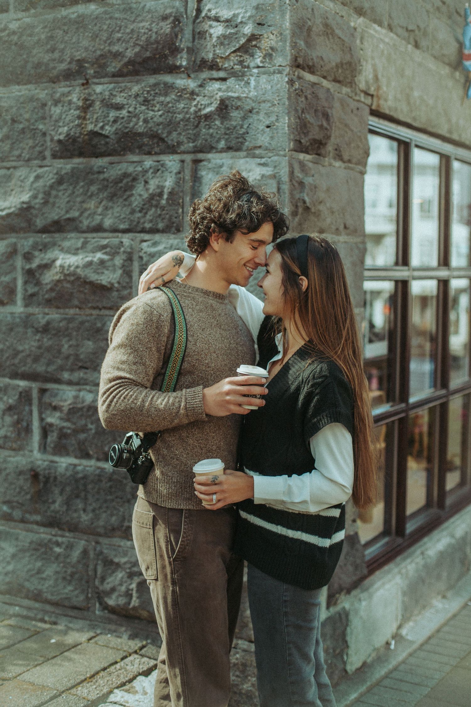 Two people in cozy sweaters share coffee and romantic kisses against a stone wall backdrop.