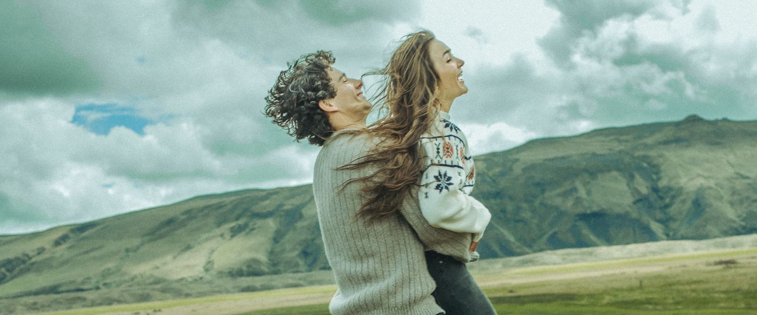 Two people share a romantic moment in a verdant mountain valley under dramatic clouds.