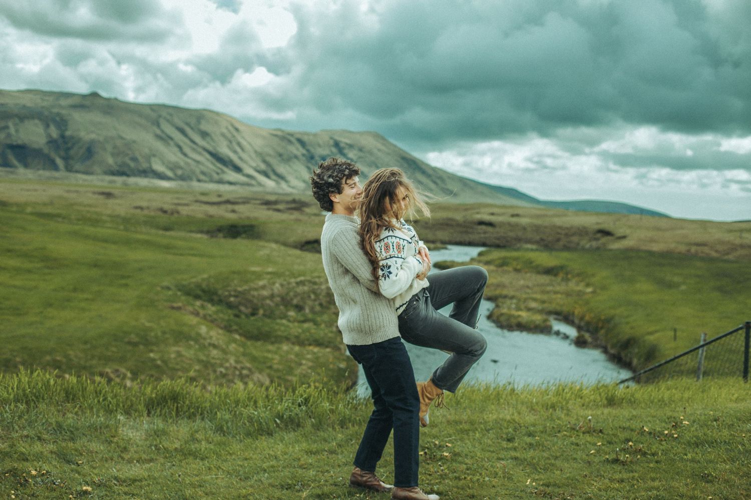 Couple embracing on hilltop with winding river and mountains in dramatic landscape while wearing cozy sweaters..