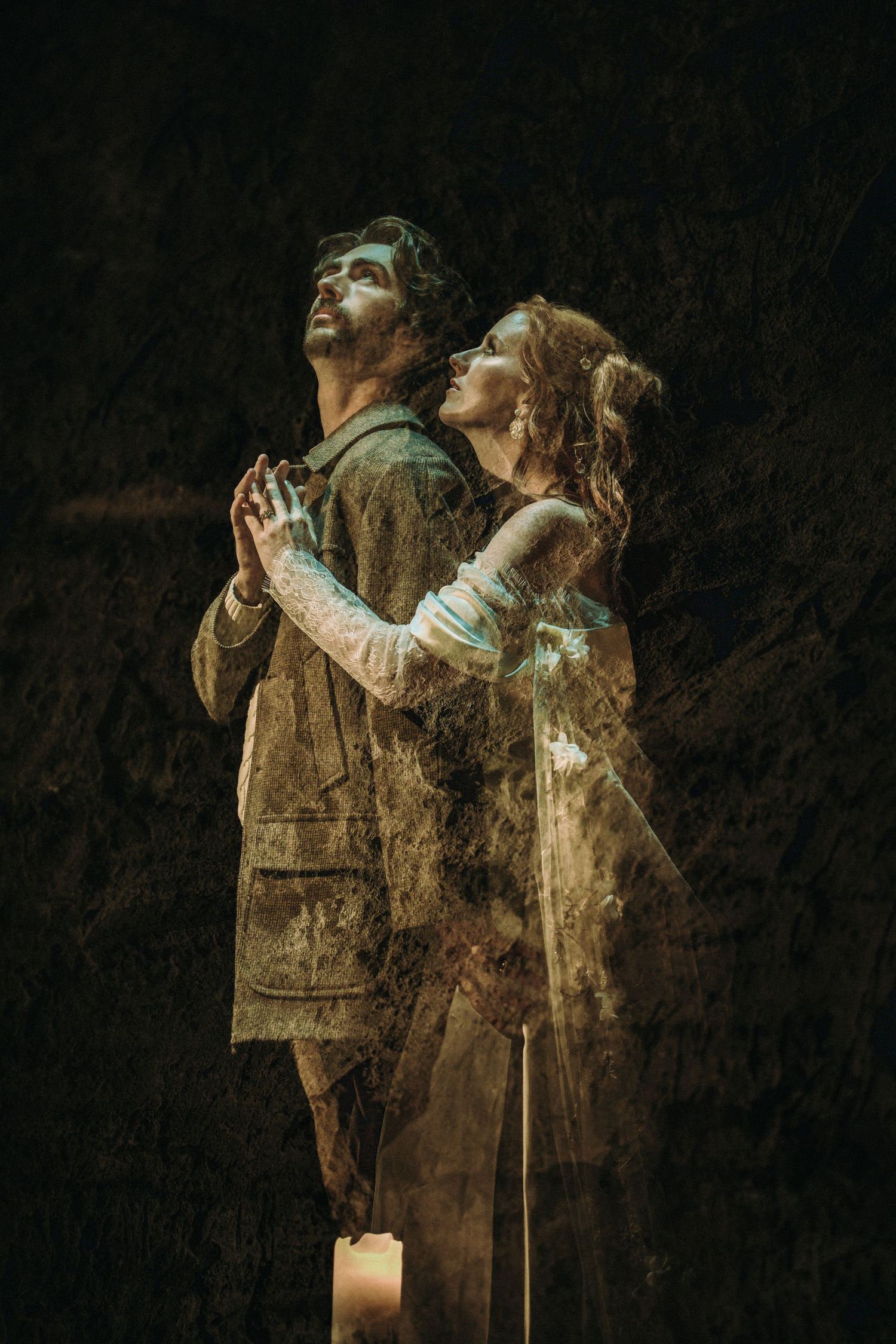 Two figures in vintage clothing stand back-to-back while gazing upward with joined hands near a glowing candle in dark ambiance.