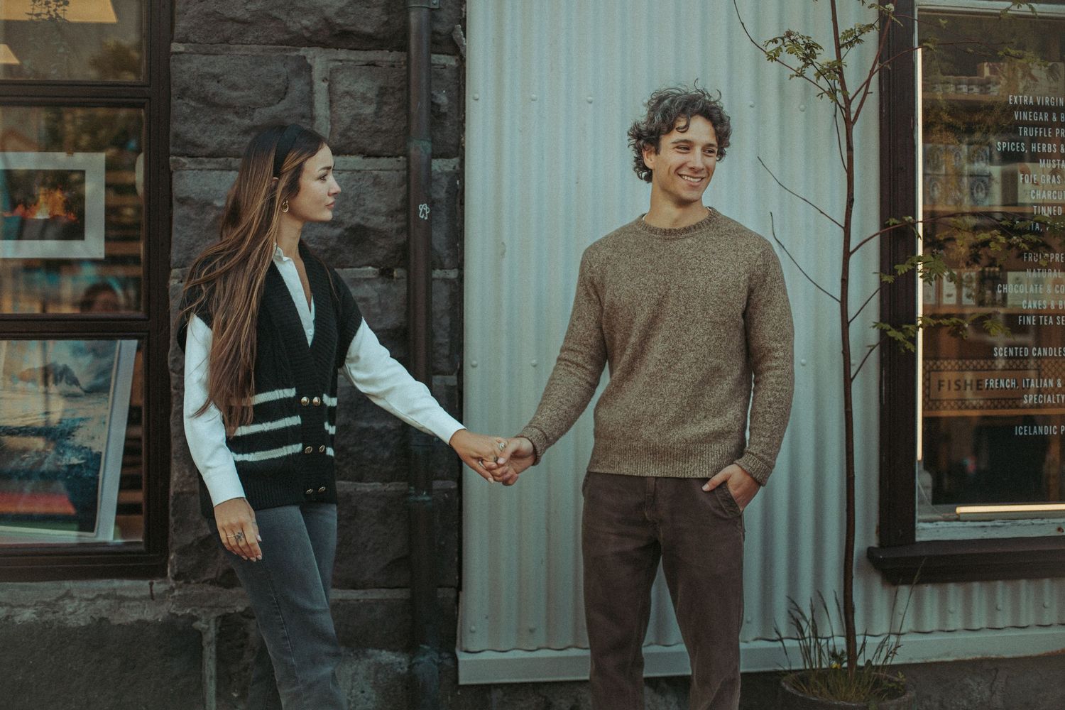 A couple holds hands while standing outside a storefront in casual autumn attire.