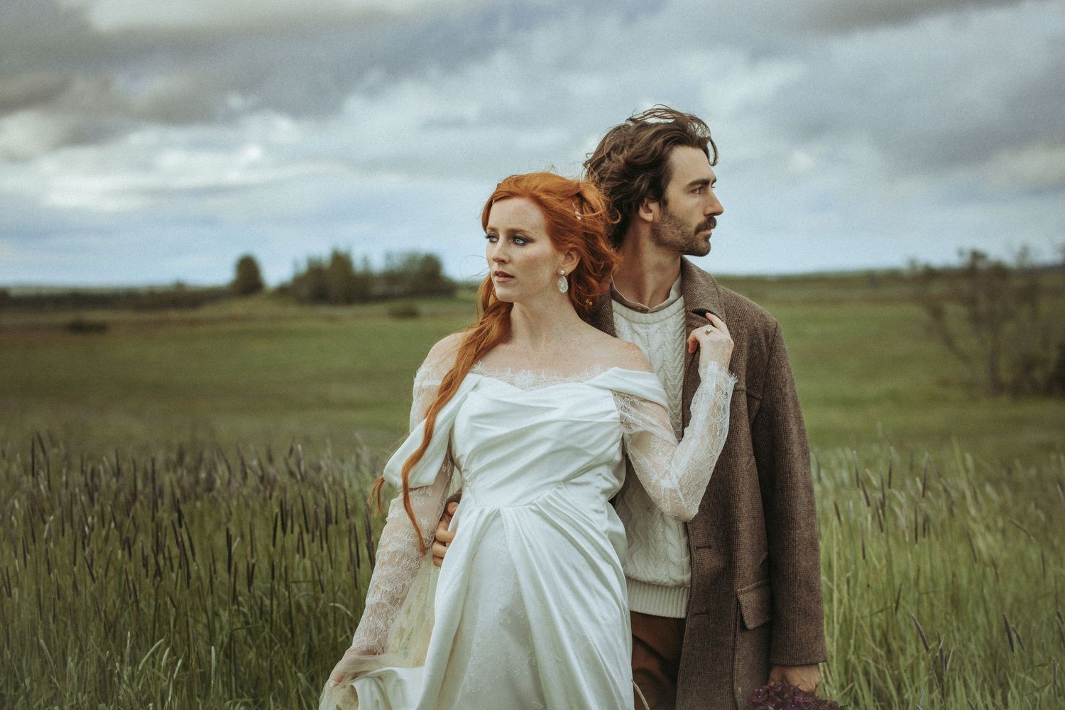 Romantic newlywed couple embrace in scenic field under moody cloudy sky during rustic countryside wedding shoot.