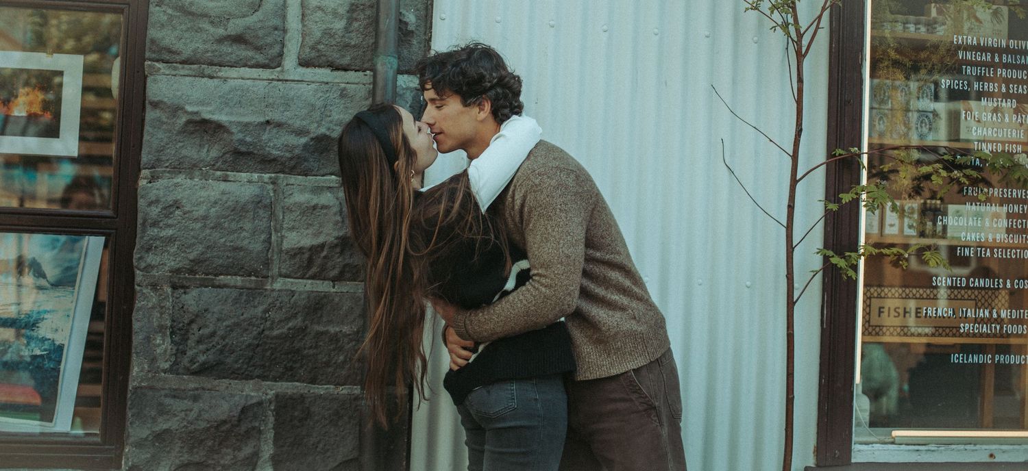 Two people in vintage-style clothing share an intimate embrace against a stone wall with some greenery visible.