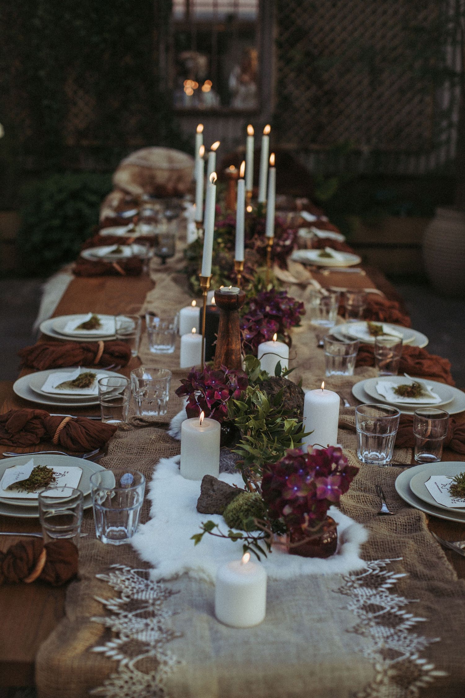 Elegant rustic table setting with white candles, purple flowers, and vintage place settings on burlap runner.