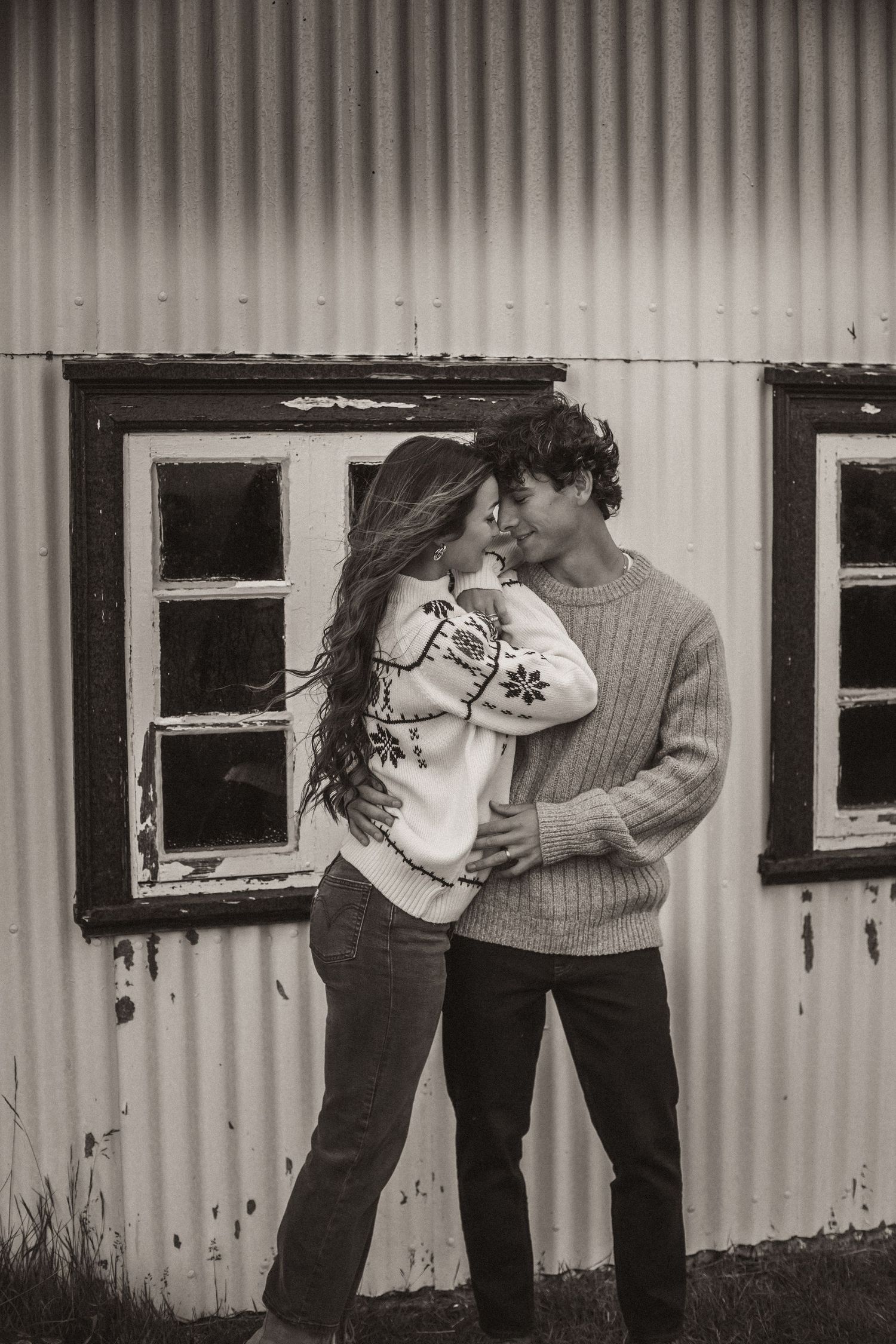 Black and white image of a couple in winter sweaters sharing an intimate moment by a rustic wall.