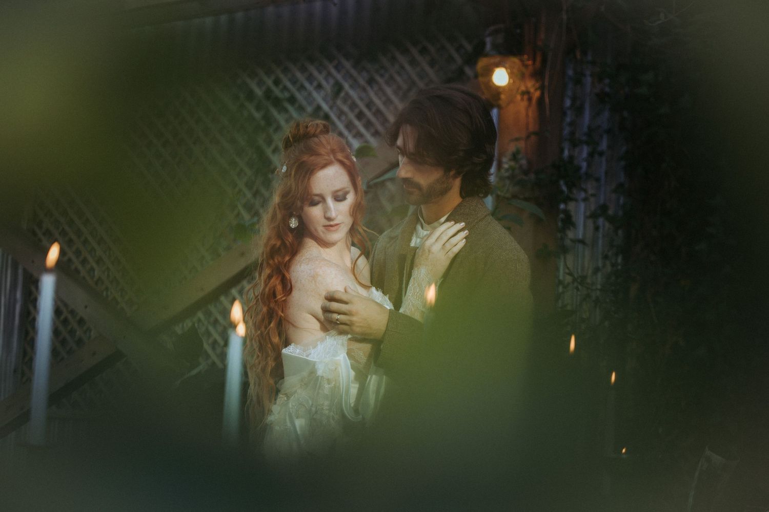 A romantic vintage-style couple in period dress share an intimate candlelit moment through blurred greenery.