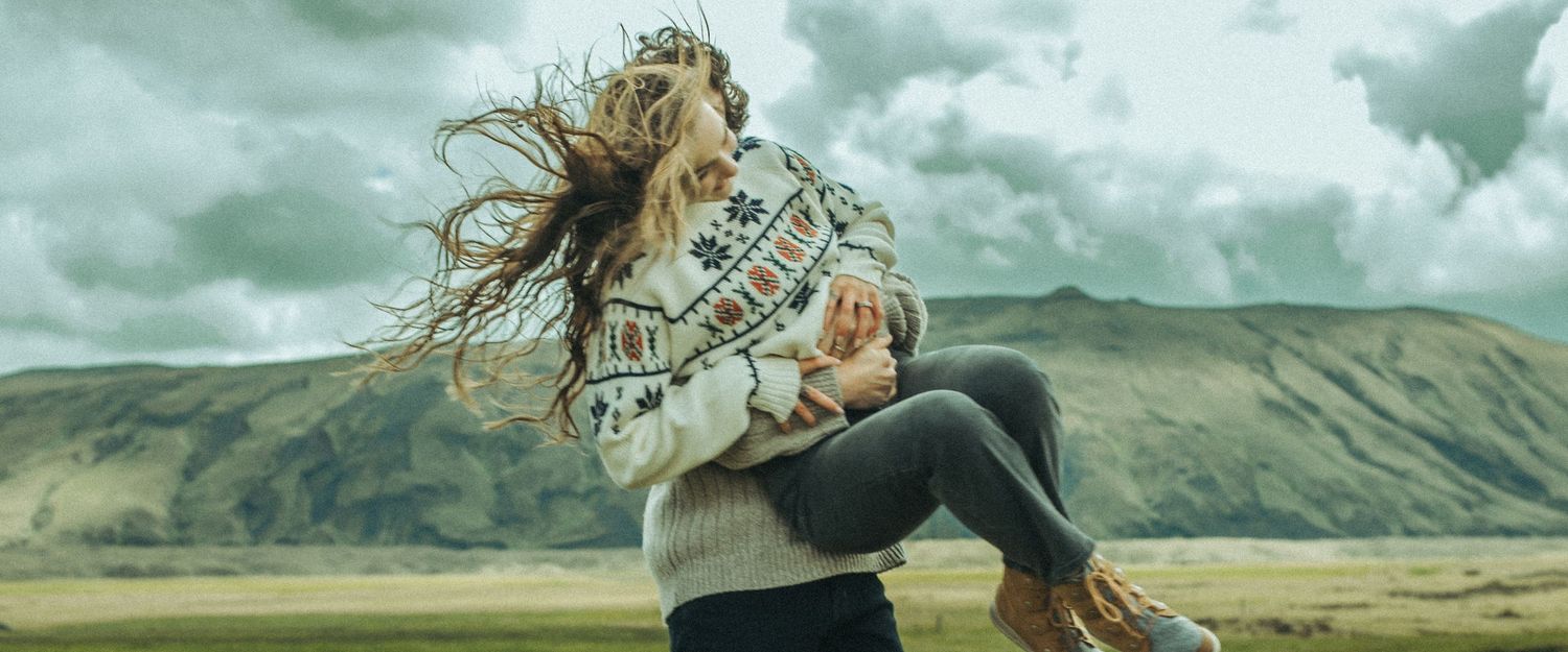 A scenic panoramic shot captures two people embracing against green mountains and cloudy skies.