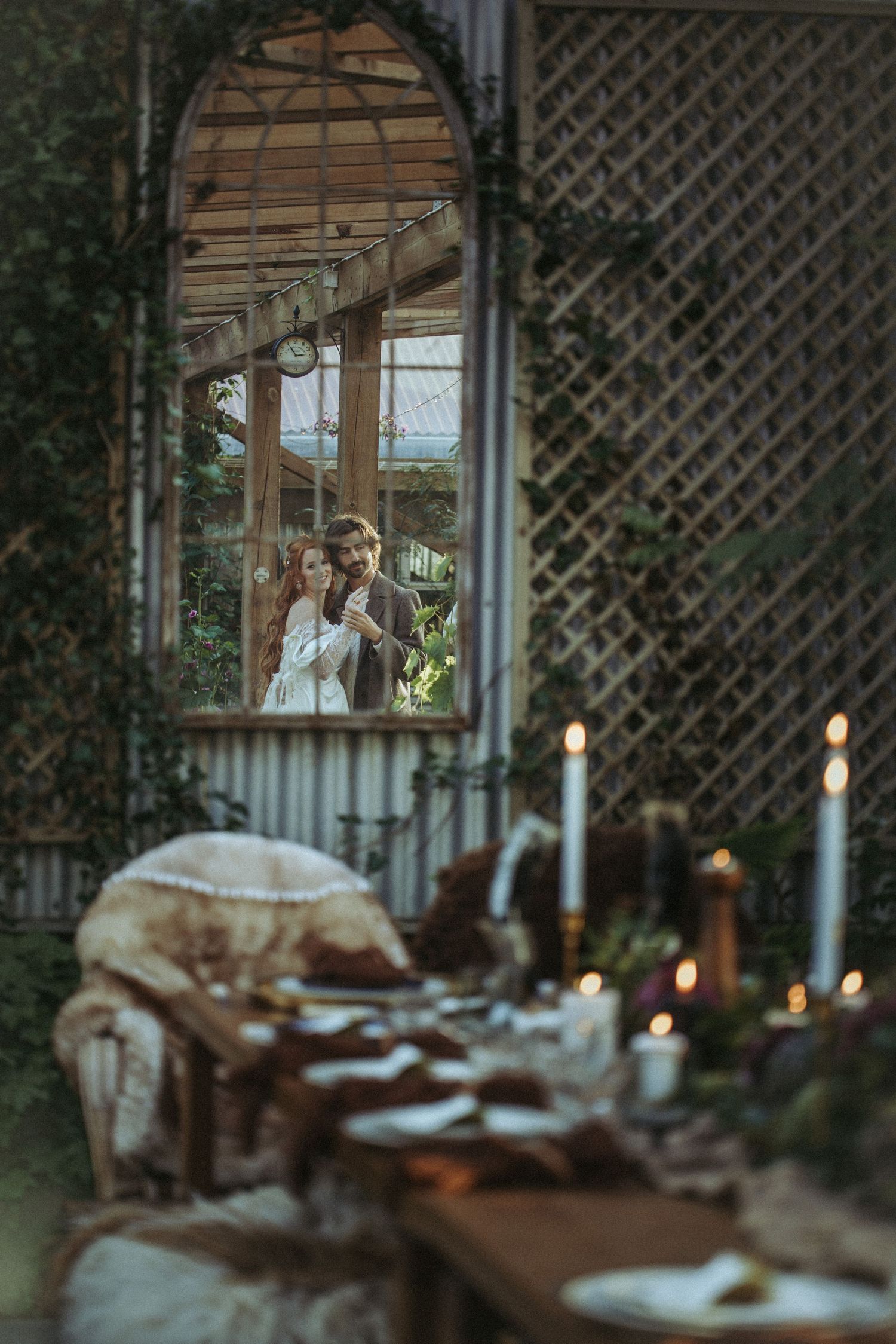 A rustic outdoor dining setup with lit candles on a wooden table, seen through a decorative lattice and window frame.