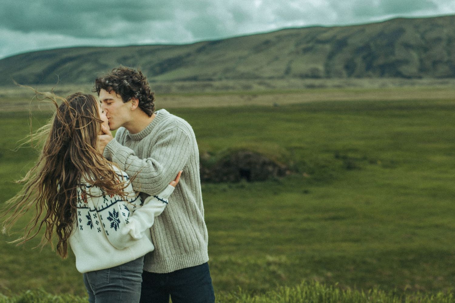 Romantic moment captured in sweeping mountain landscape with dramatic clouds while wearing cozy sweaters..