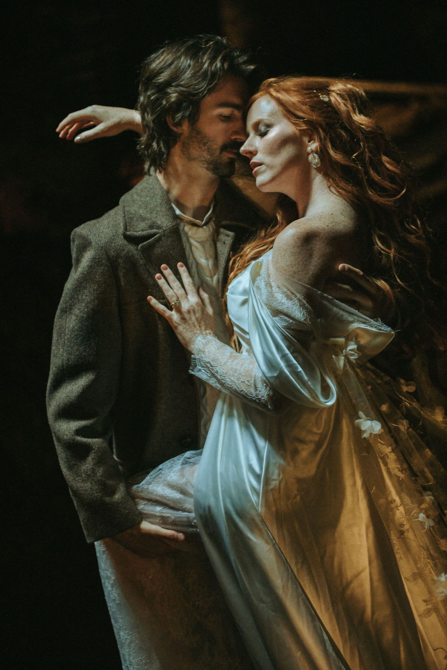 A romantic vintage-style portrait of a couple embracing, with the woman wearing a flowing white dress in moody lighting.