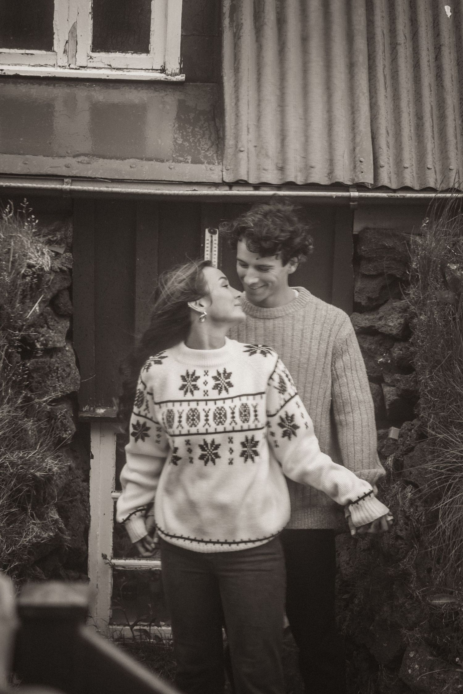 Couple in matching winter sweaters sharing a laugh outside a rustic building.