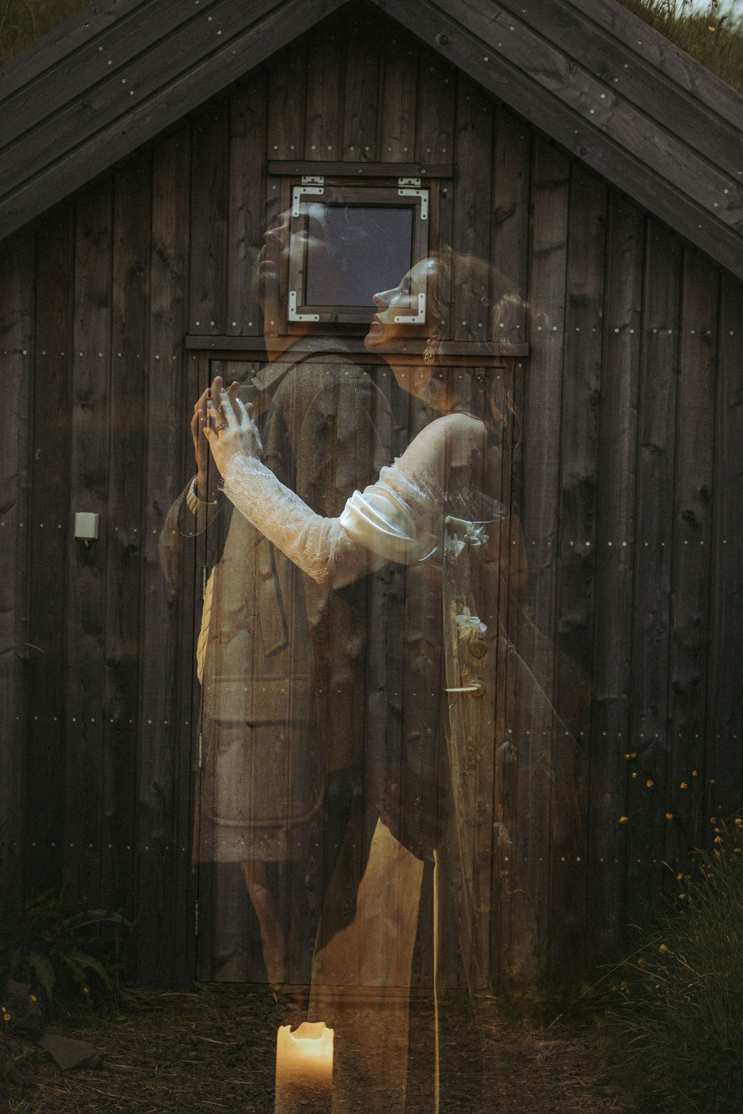 A ghostly ethereal figure stands in front of a dark wooden cabin with a small lit candle on the ground.