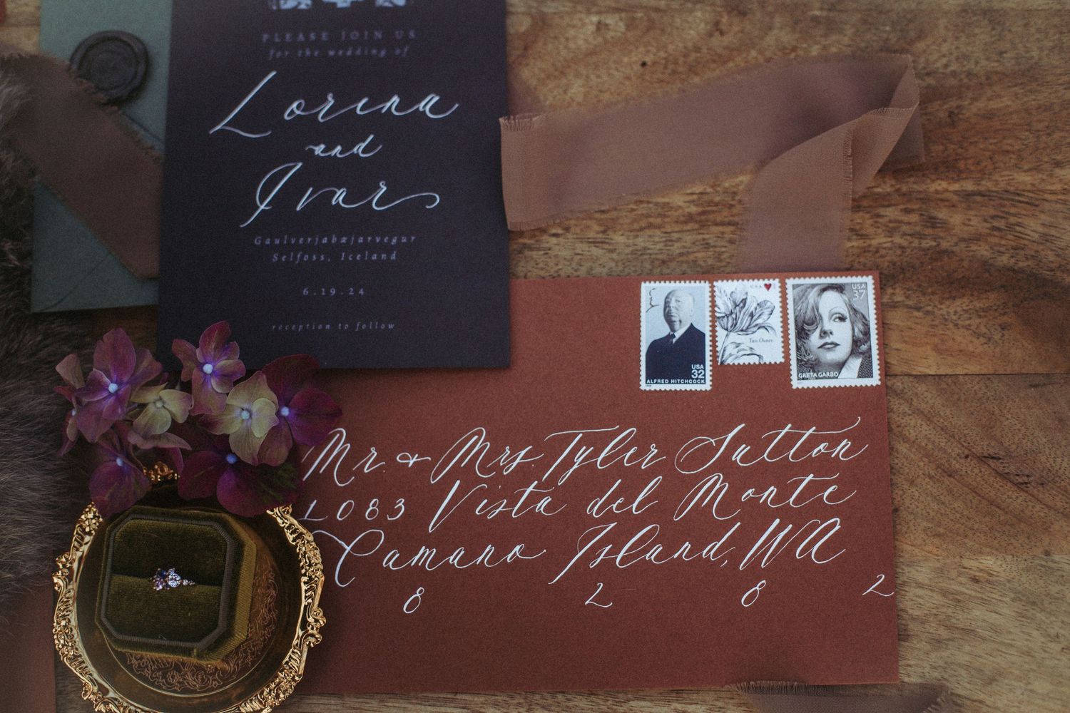 Wedding invitation suite with calligraphy on burgundy envelope, vintage stamps, and dried flowers on wooden surface.