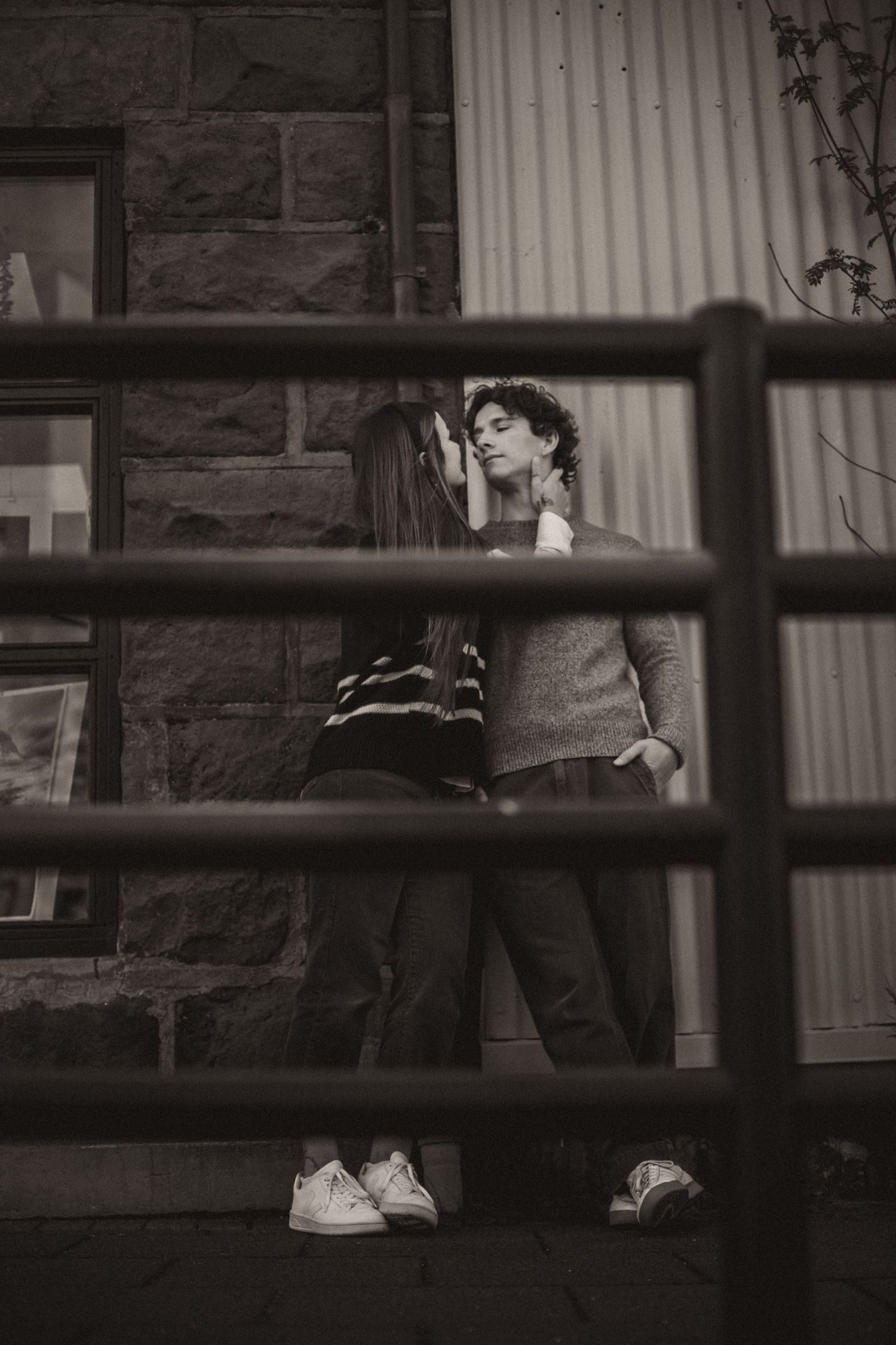 A black and white photo shows two people sitting on a bench through wooden fence slats.