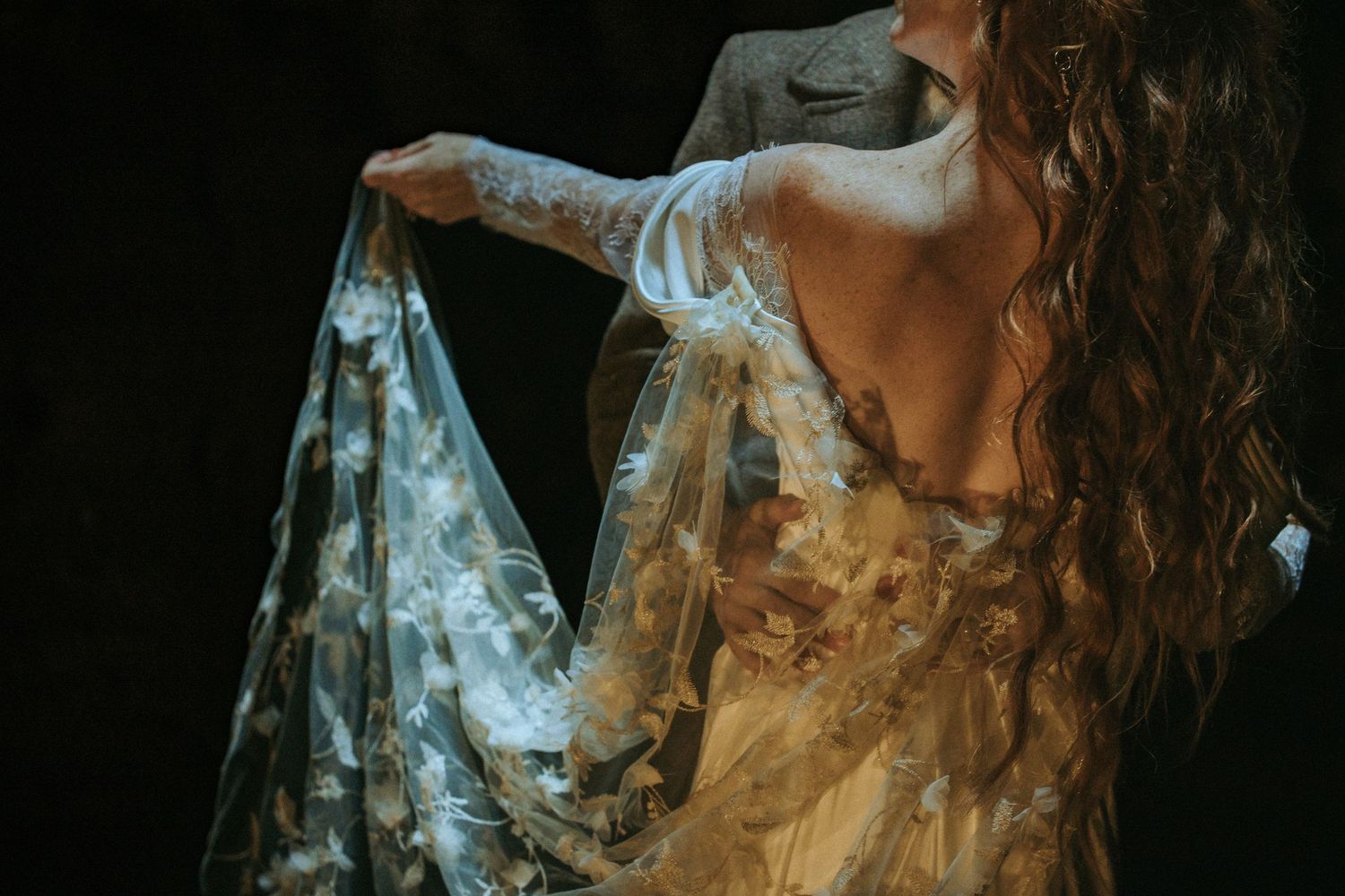 A ethereal close-up shot shows someone holding a delicate floral sheer fabric against a dark background.