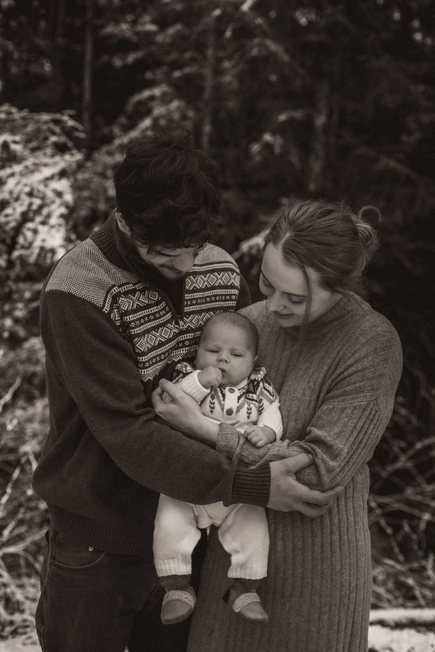 A tender family moment in the snowy forest showcasing parents holding their baby in warm winter sweaters.