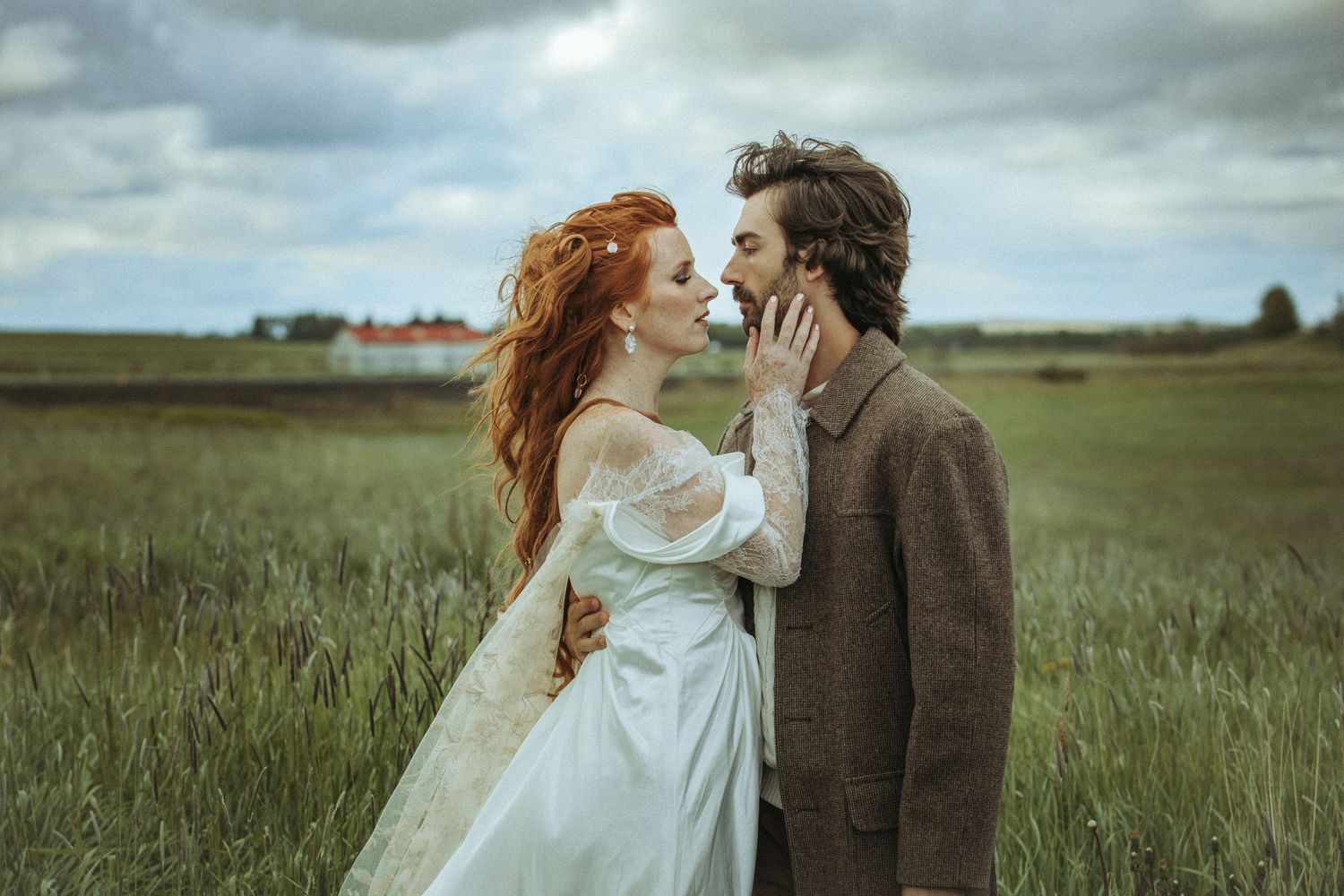 A romantic couple stands together in a windswept field against a cloudy sky with a vintage-inspired aesthetic.