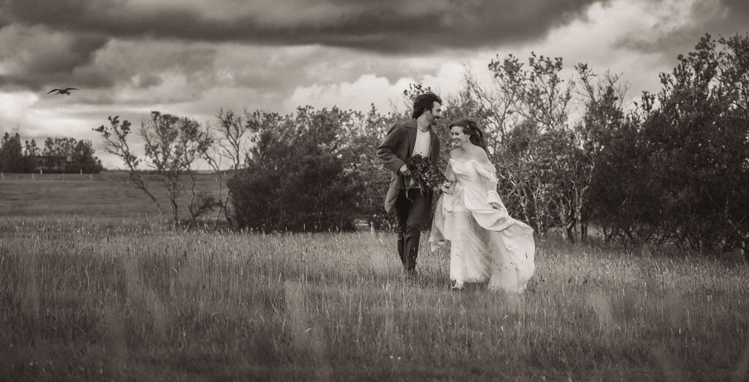 Dramatic black and white photograph of couple posing in a prairie field under stormy clouds at sunset.