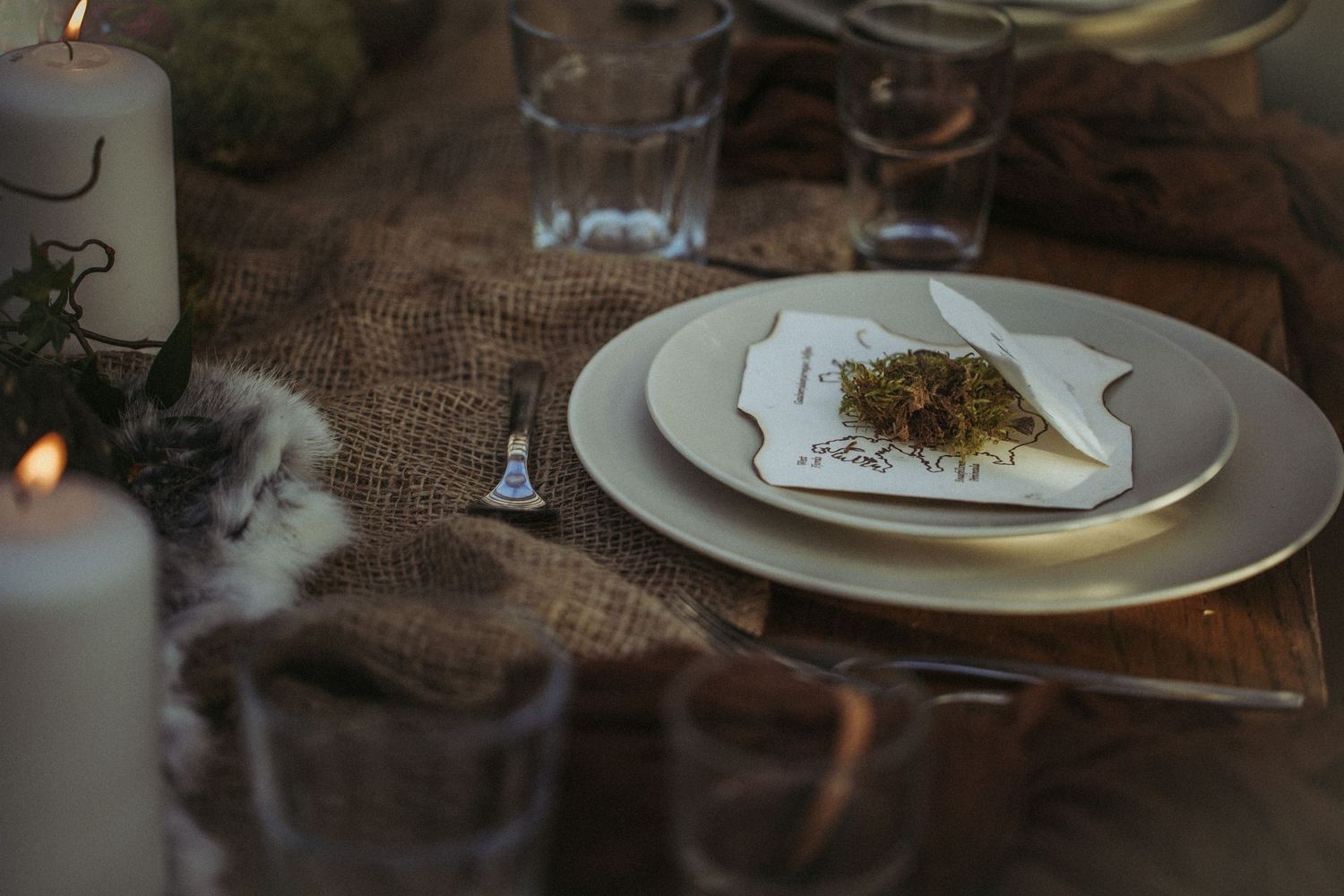 A rustic dining table setting with white plates and a lit candle on burlap cloth in moody lighting.