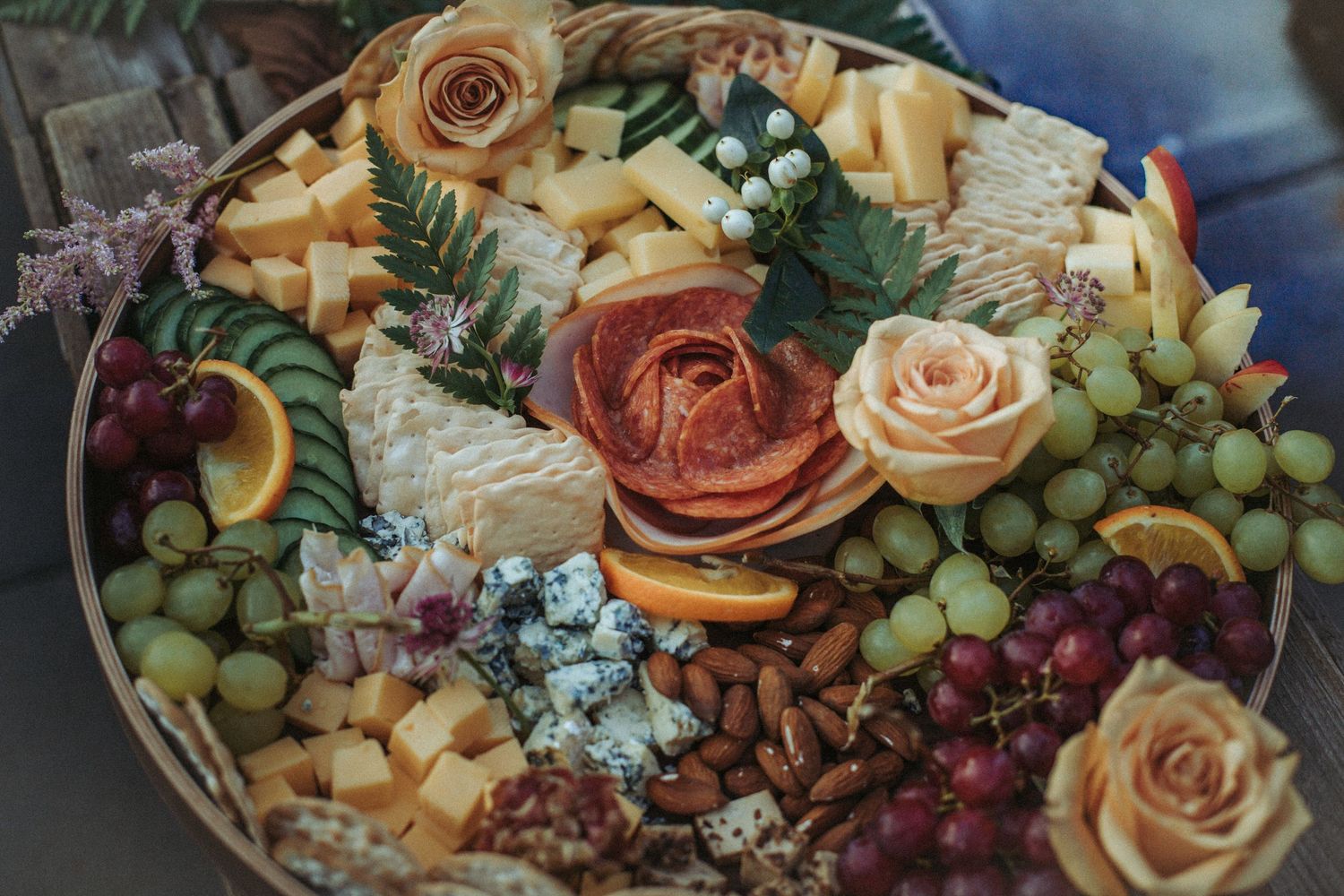 A gourmet cheese and fruit platter with roses, grapes, citrus slices and blue cheese arranged on a decorative board.