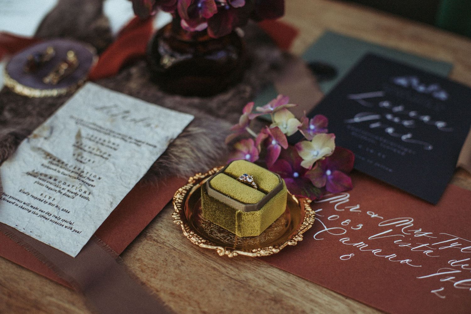 A golden dessert on a small plate surrounded by purple flowers and invitation cards on a wooden surface.
