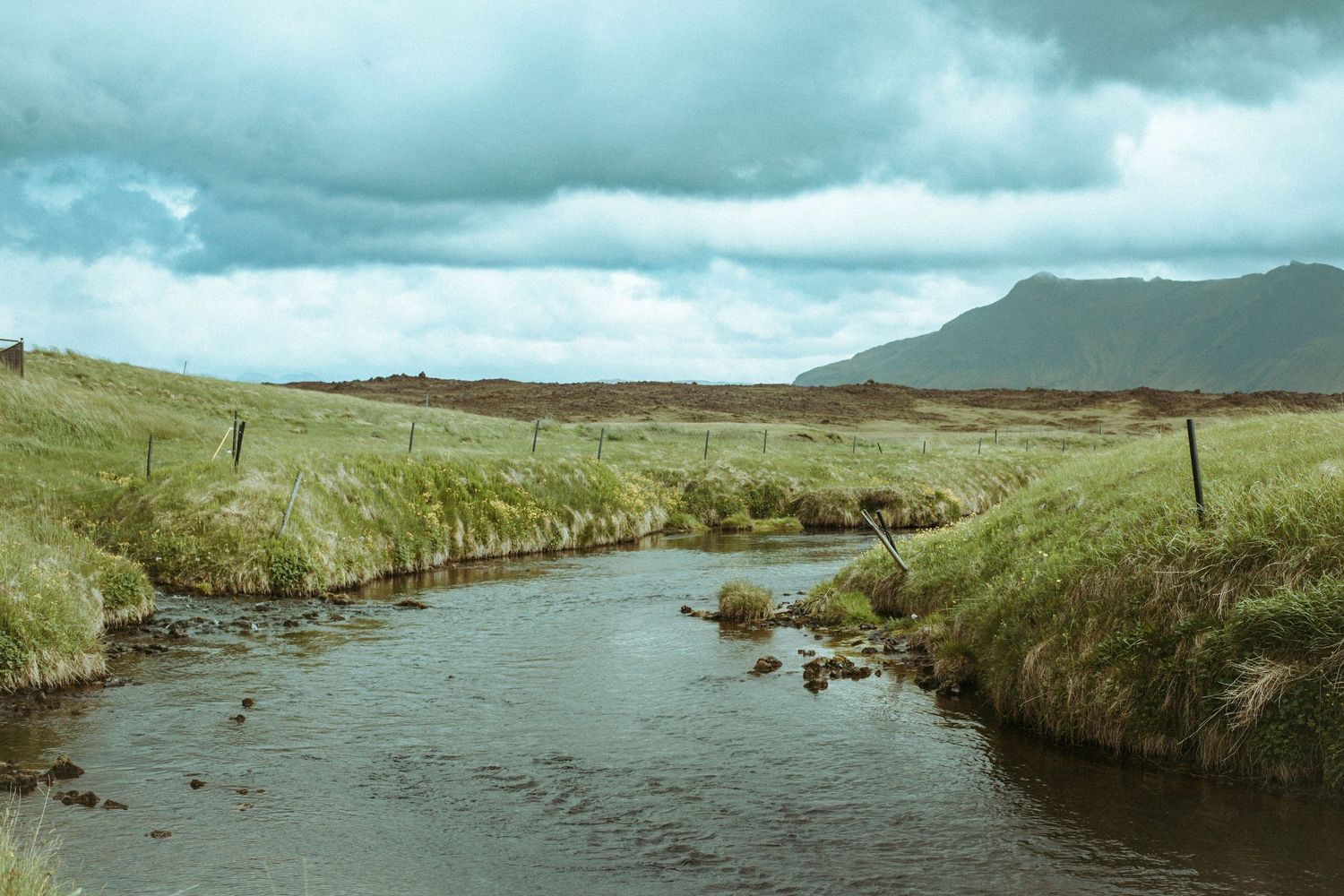 Winding river flows through green grassland with mountains and stormy clouds in the background.