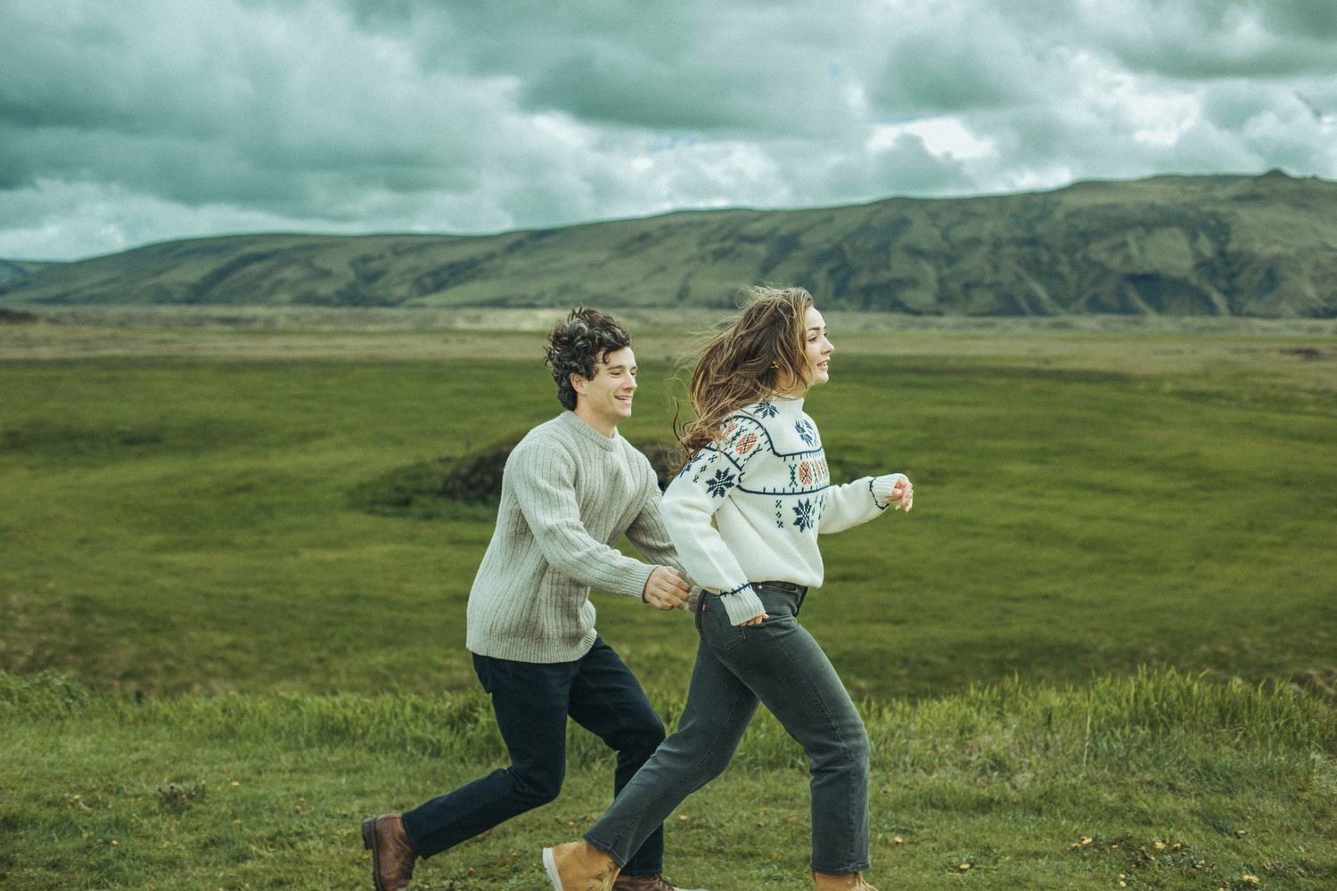 Two people running through scenic green hills under dramatic cloudy skies while wearing cozy sweaters.