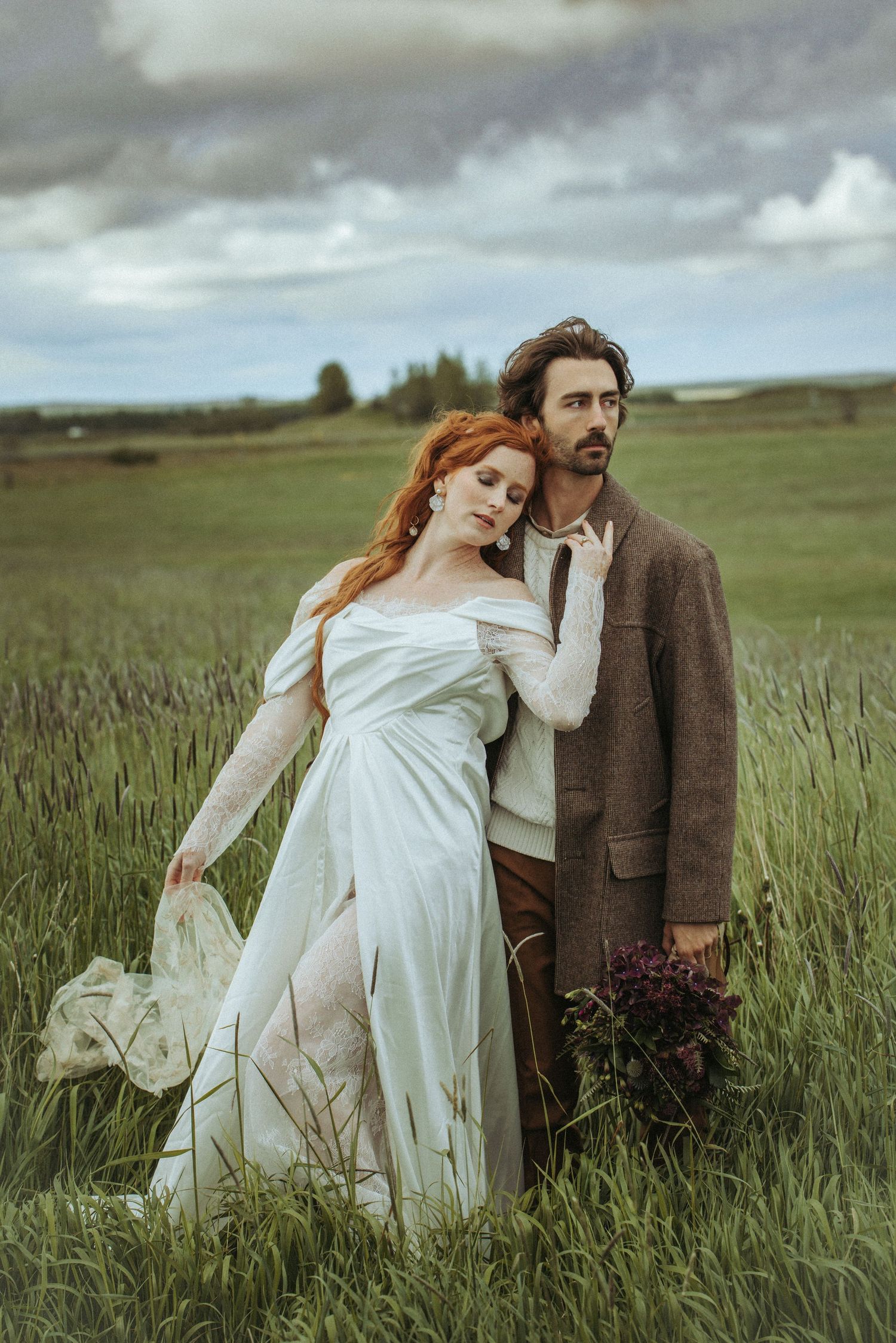 A couple in a dramatic vintage-inspired pose stands in a windswept meadow with tall grass and moody cloudy skies.
