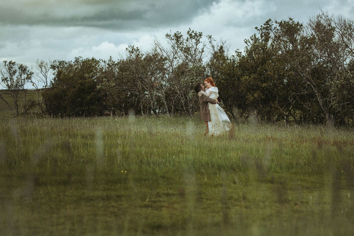 A figure in a white dress stands alone in a misty meadow surrounded by distant trees under a moody sky.