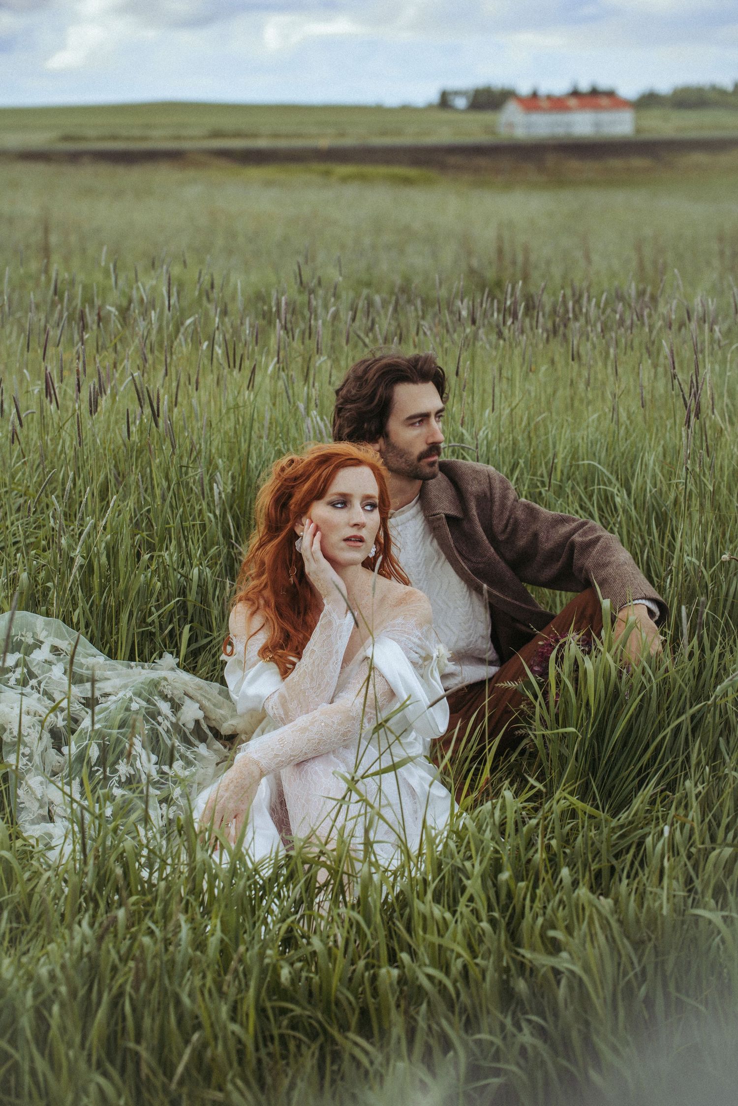 Couple lying in tall grass meadow wearing romantic vintage-inspired clothing against cloudy rural landscape.