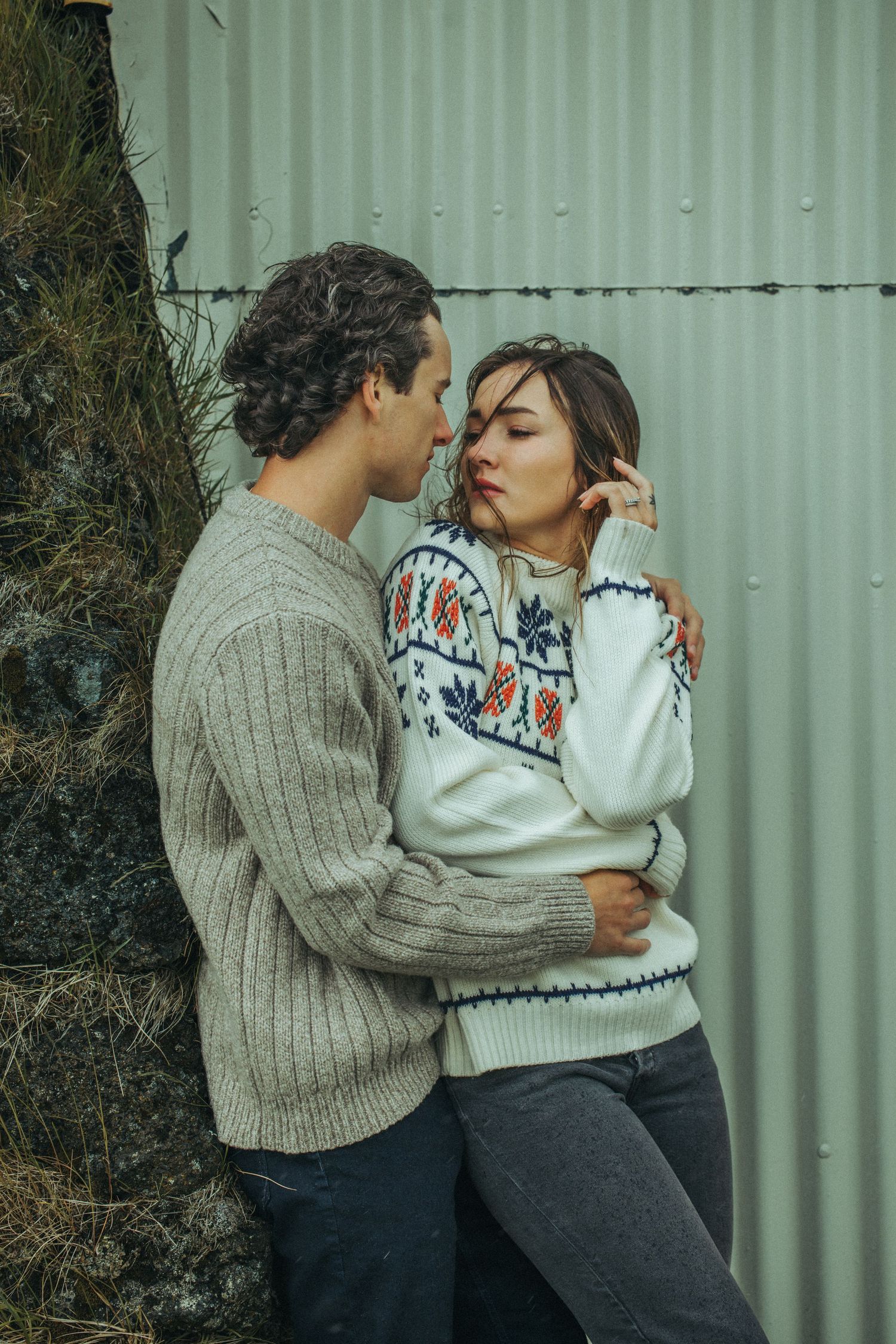 Couple in cozy winter sweaters sharing an intimate moment against a green wall outdoors.