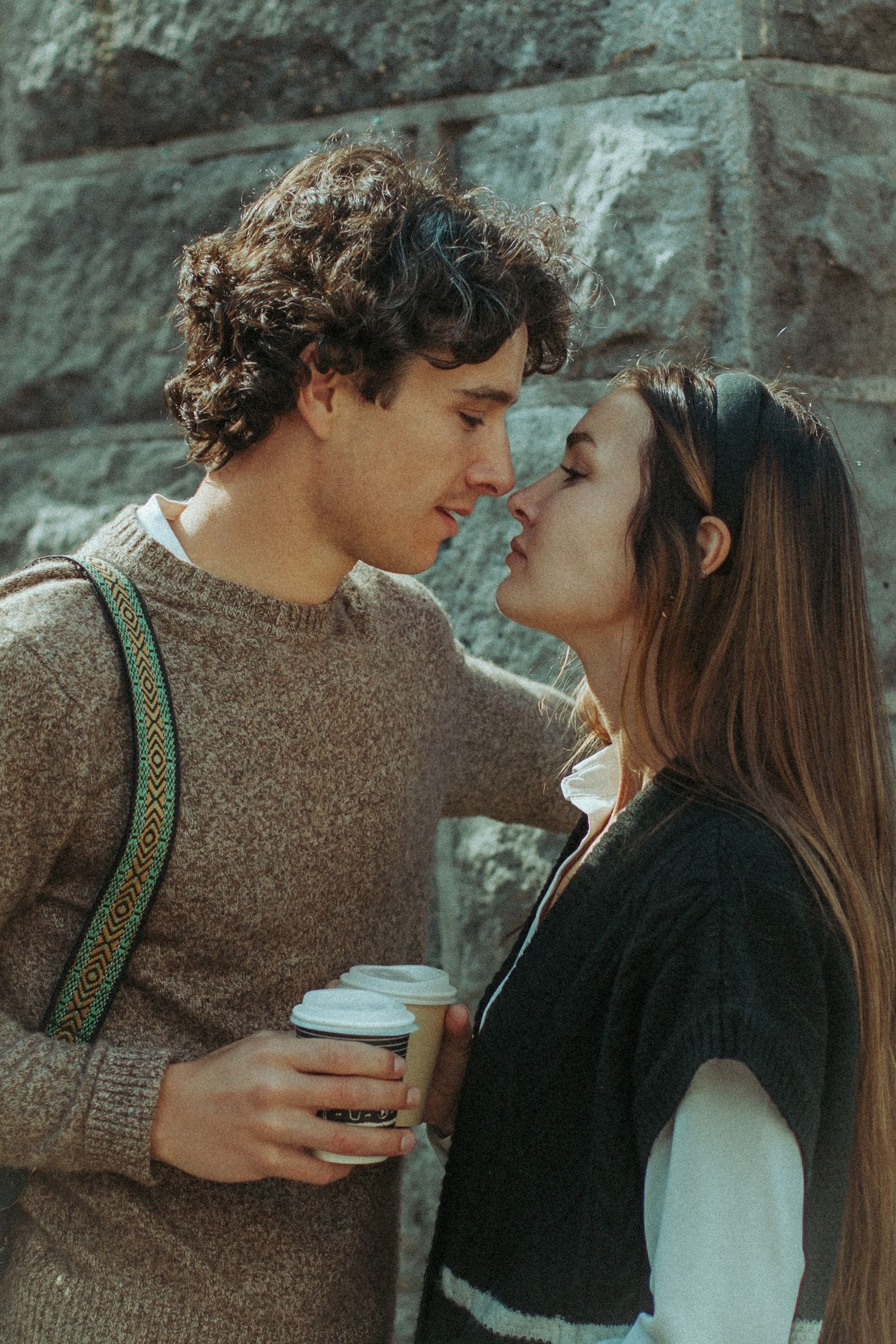 A romantic couple shares an intimate moment against a stone wall while one holds a coffee cup.